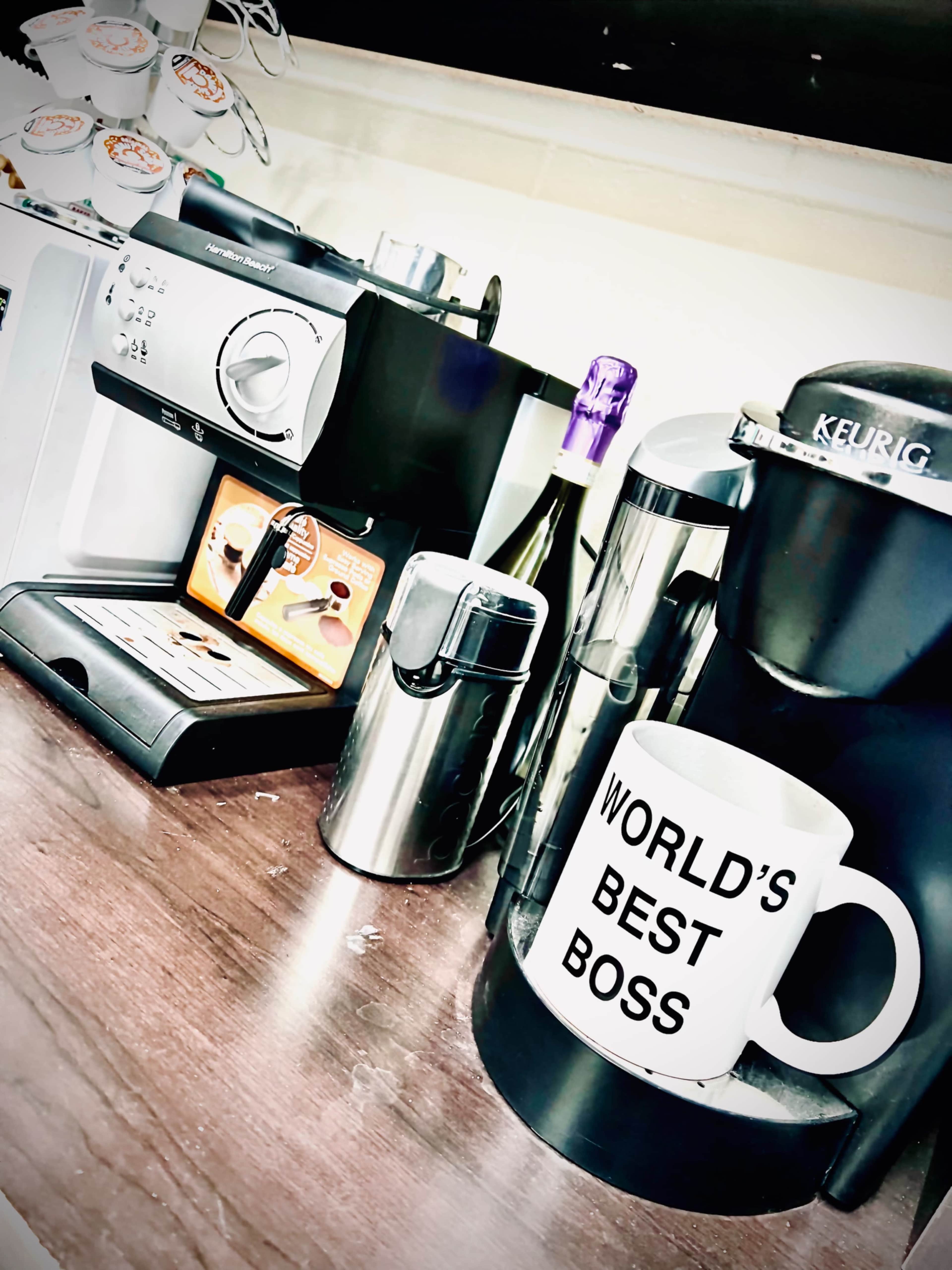 A coffee station features a Keurig machine, an espresso machine, a silver canister, and a large mug labeled "WORLD'S BEST BOSS."