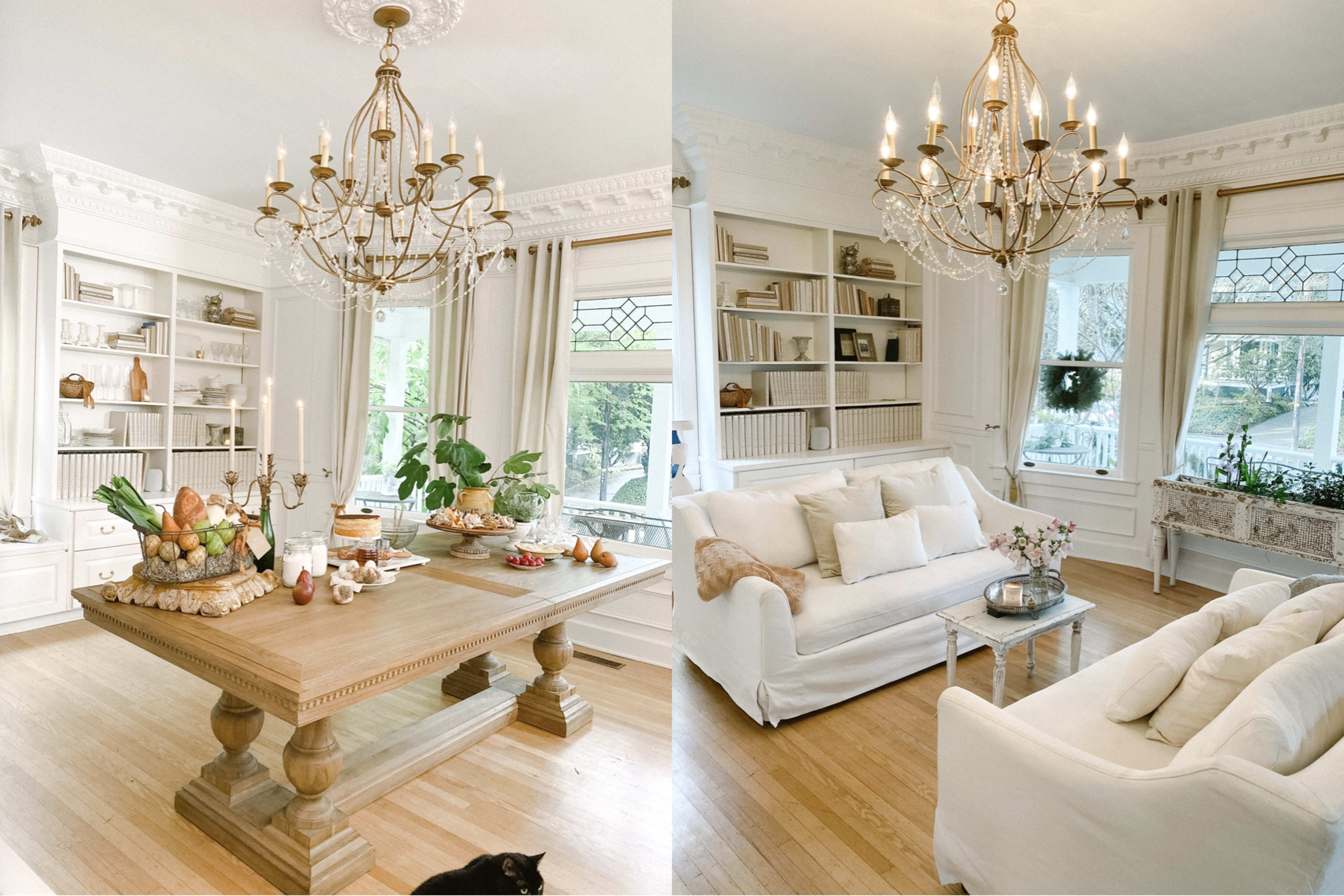 A bright living room features a wooden table set with food and candles, surrounded by white furniture and large windows letting in natural light.