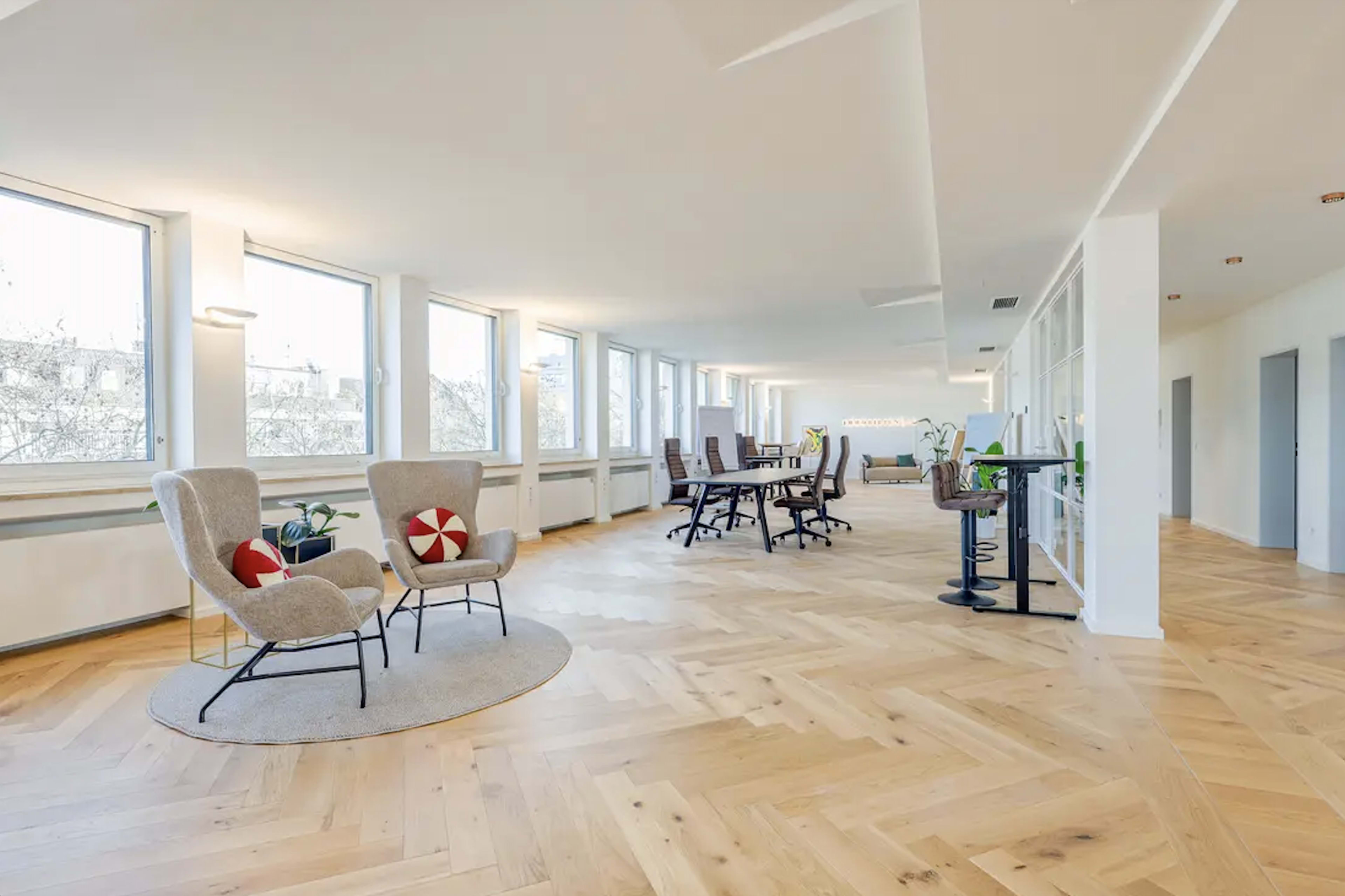The image shows a bright, spacious office area with large windows, wooden flooring, and modern furniture arranged for collaborative work and seating.
