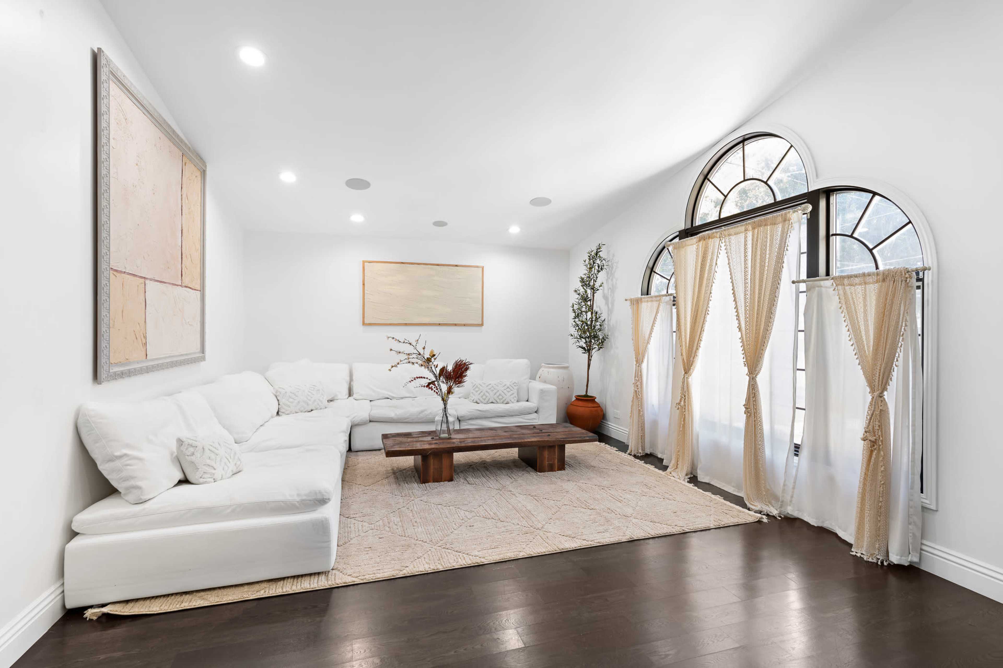 A bright living room features a large, white sectional sofa, a wooden coffee table, and arched windows draped with sheer curtains.