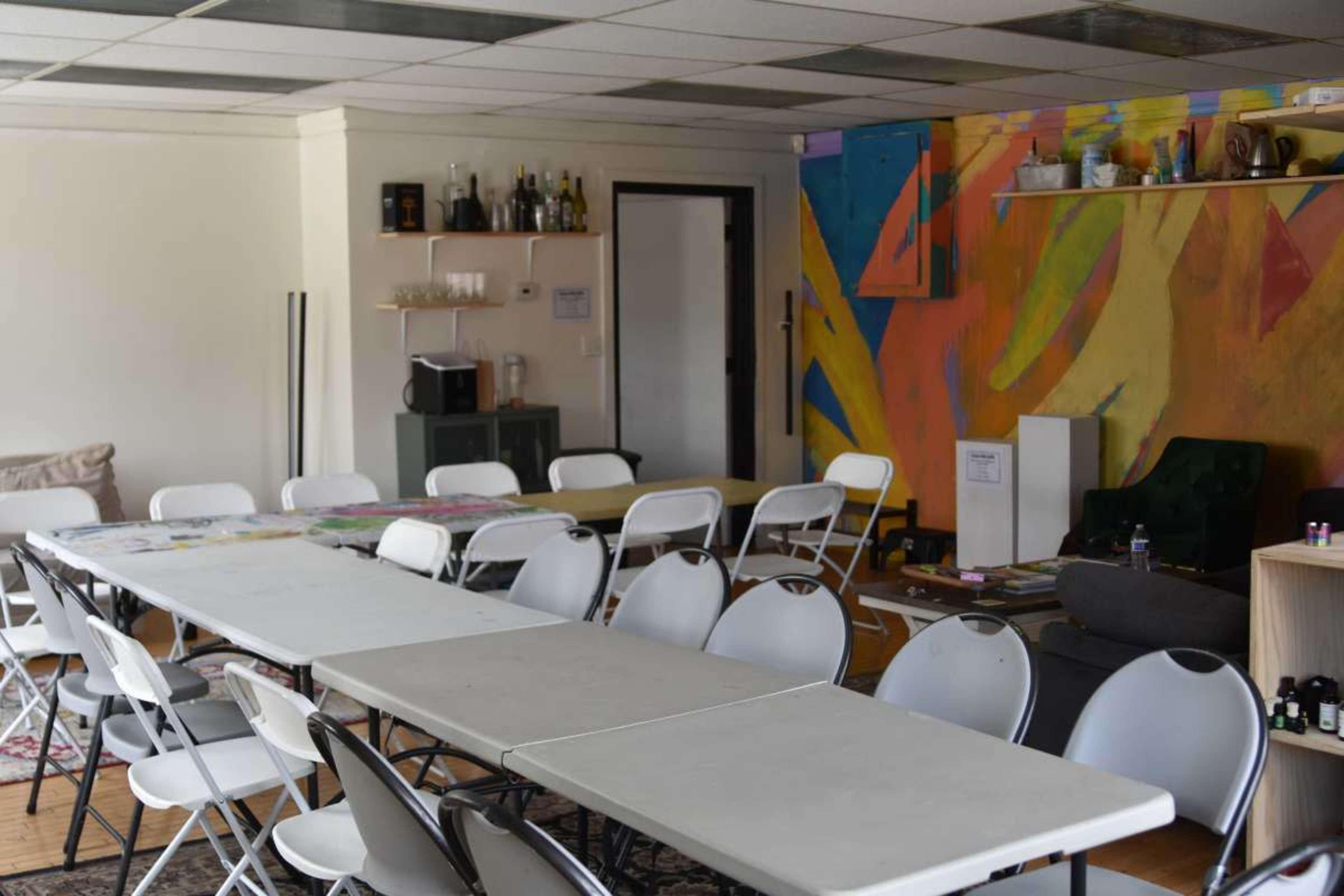The image shows a spacious room with several white folding tables and chairs arranged in rows, featuring a colorful mural on one wall and shelves with various items.
