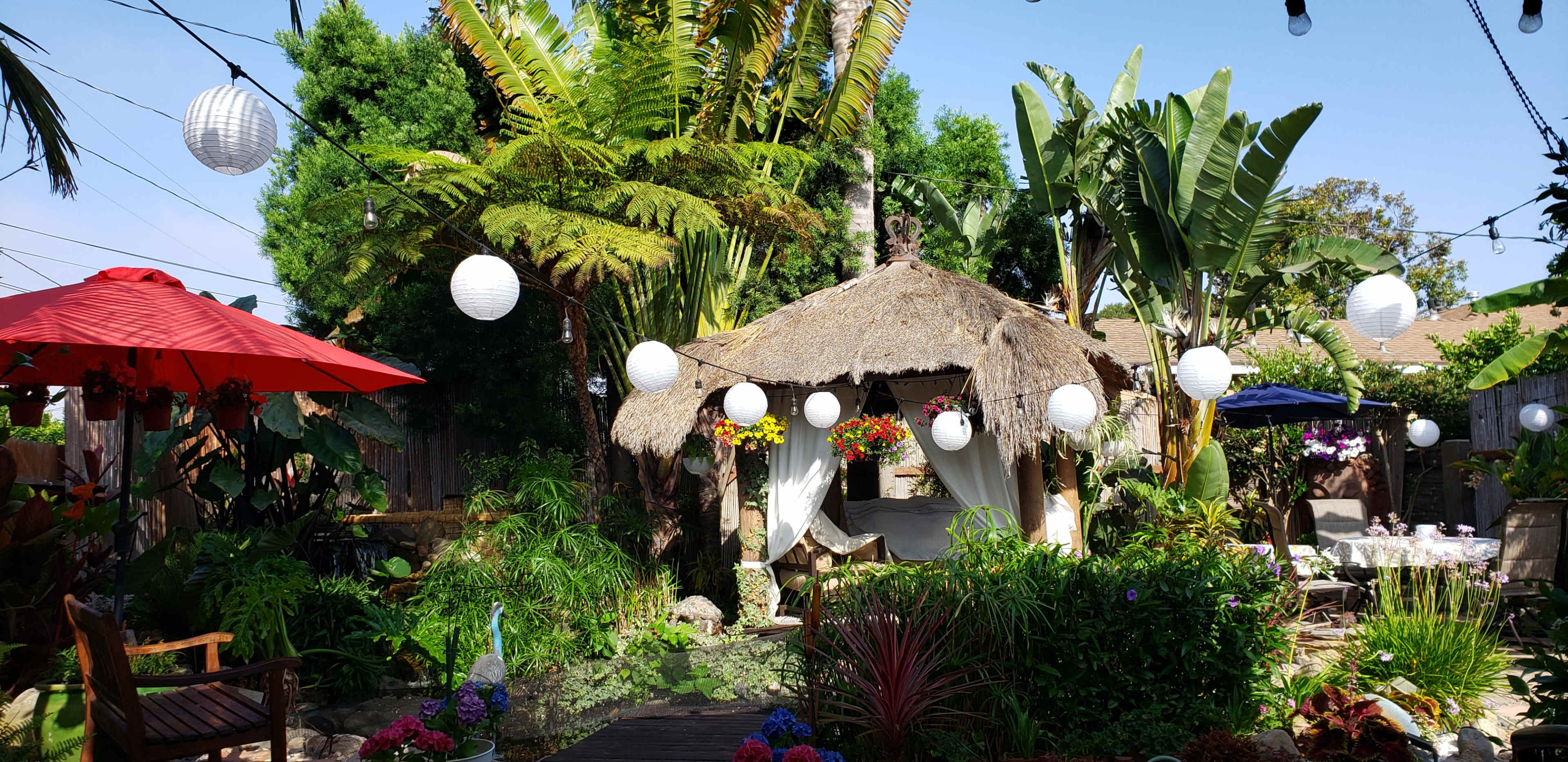 The image shows a vibrant garden with a thatched-roof gazebo surrounded by tropical plants and hanging lanterns.