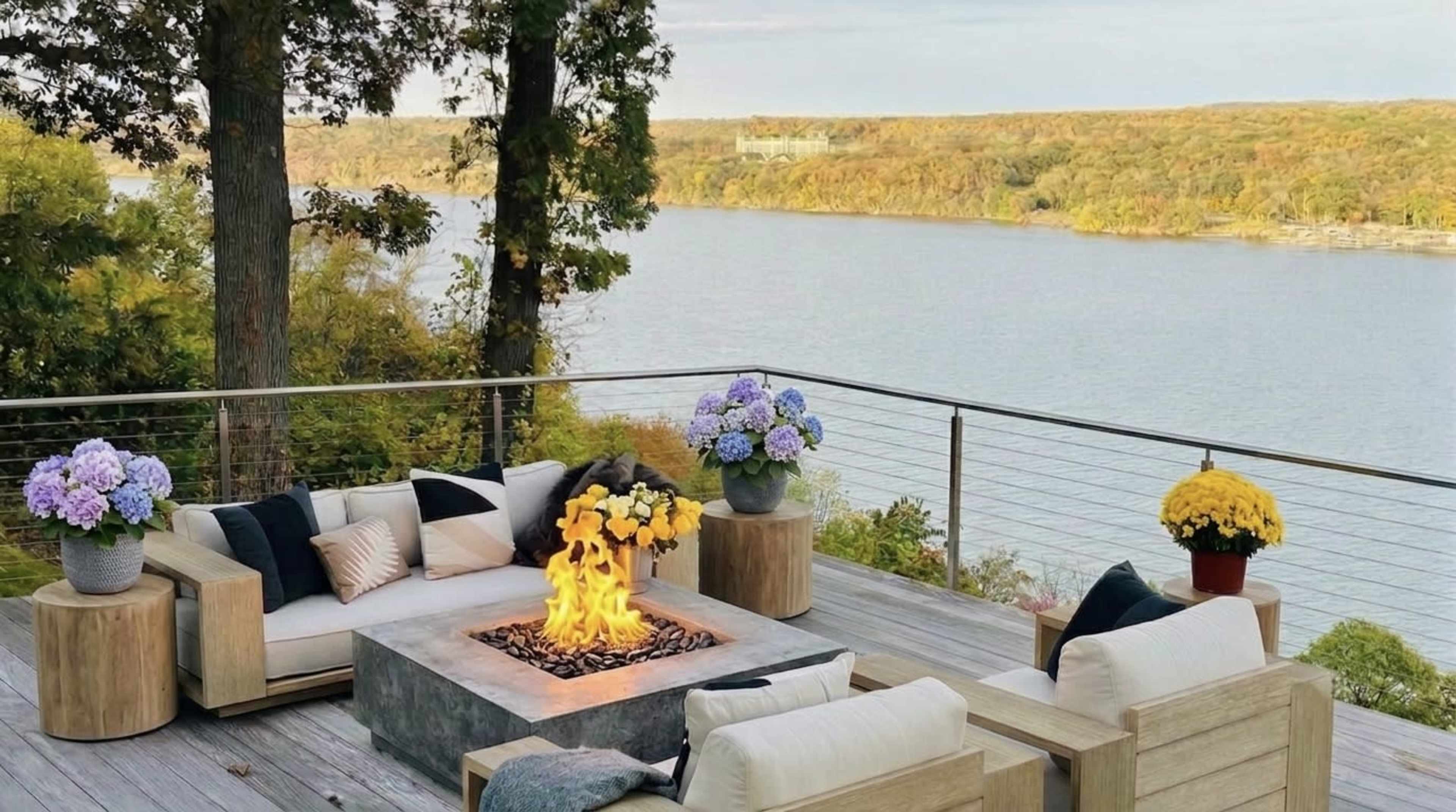 A modern outdoor seating area features a fire pit surrounded by wooden furniture and potted flowers, overlooking a body of water and wooded landscape.
