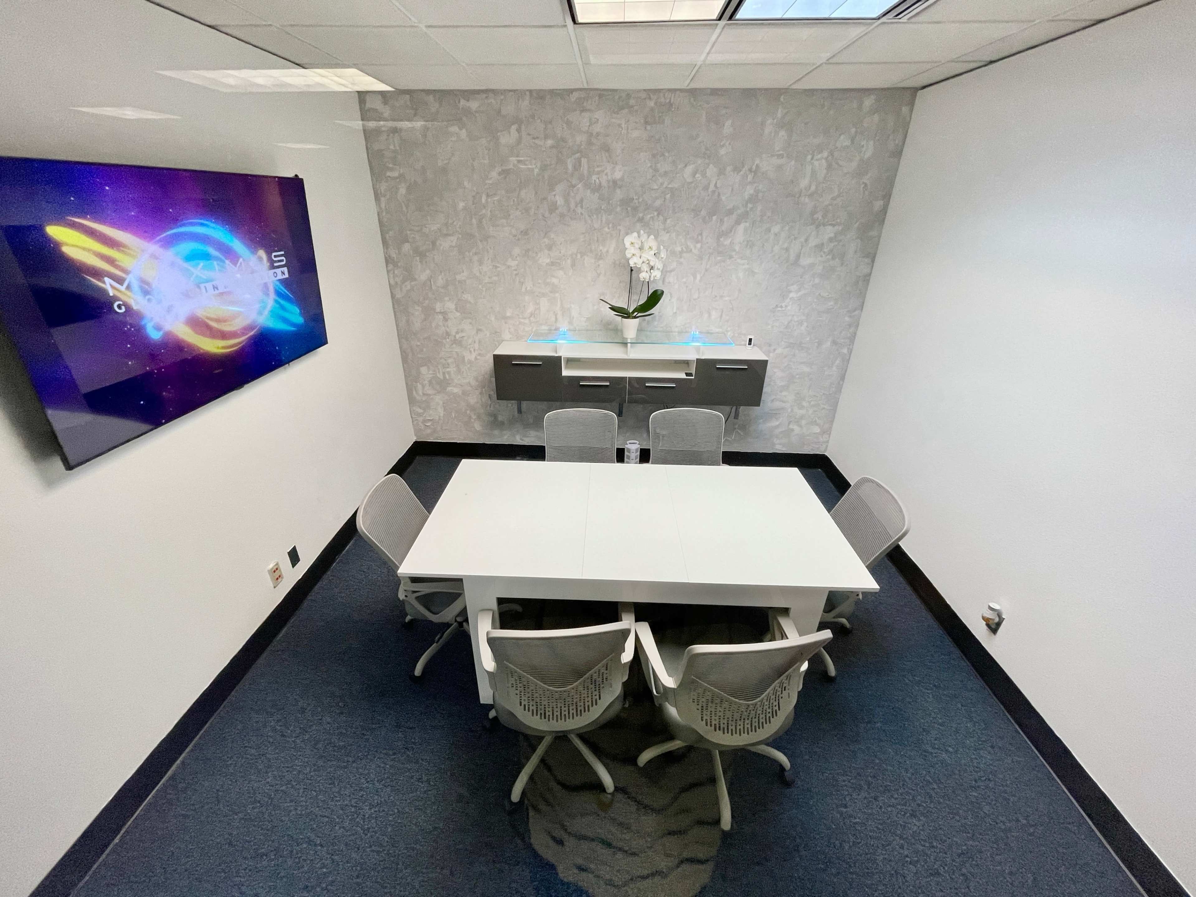 A conference room features a white table surrounded by six chairs, a mounted television, and a minimalist wall décor with an orchid centerpiece.