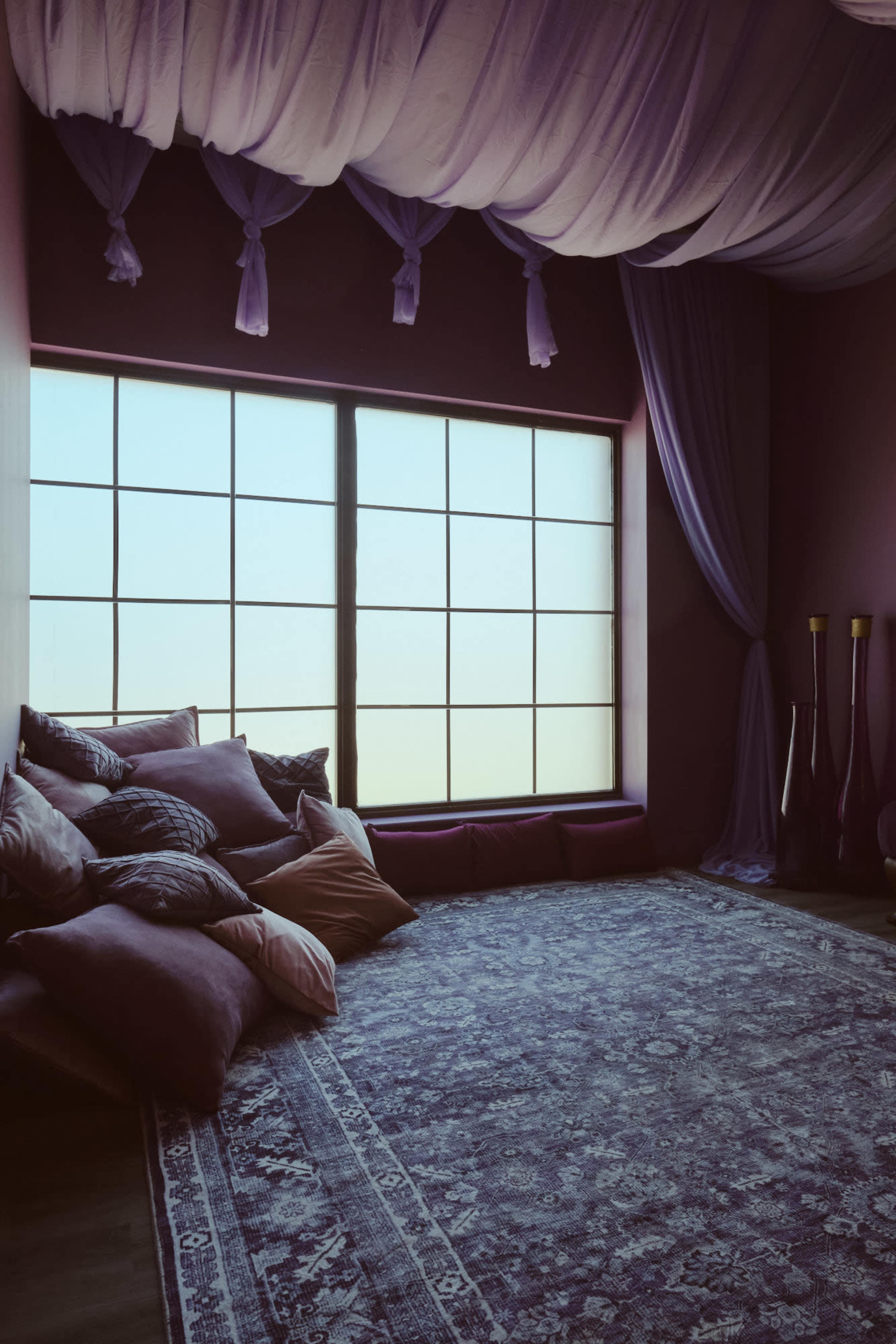 The image features a softly-lit room with a large window, plush cushions scattered on the floor, and hanging draped fabric overhead.