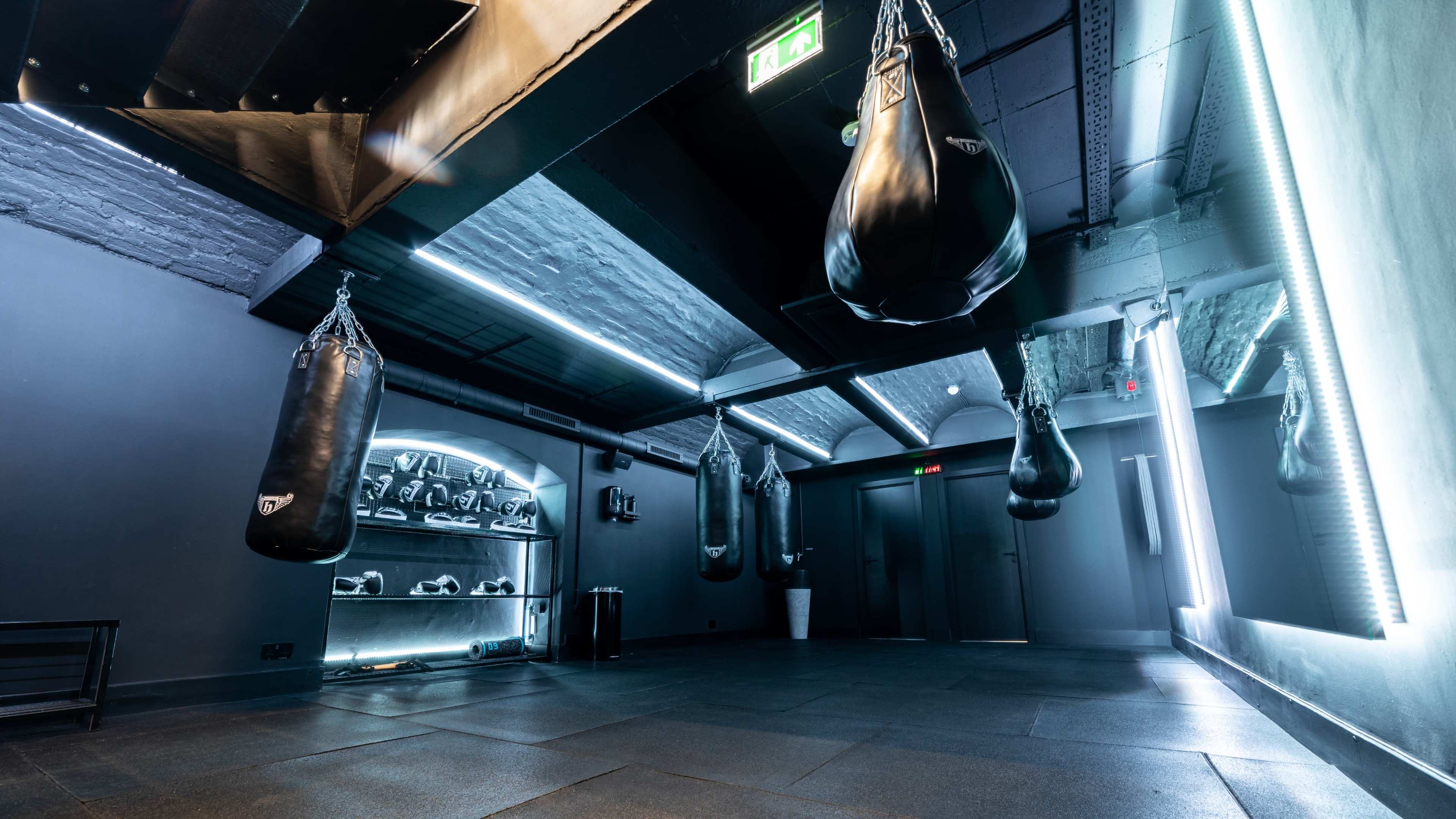 The image shows an empty boxing gym with several hanging punching bags and a display of boxing gear on a backlit shelf.