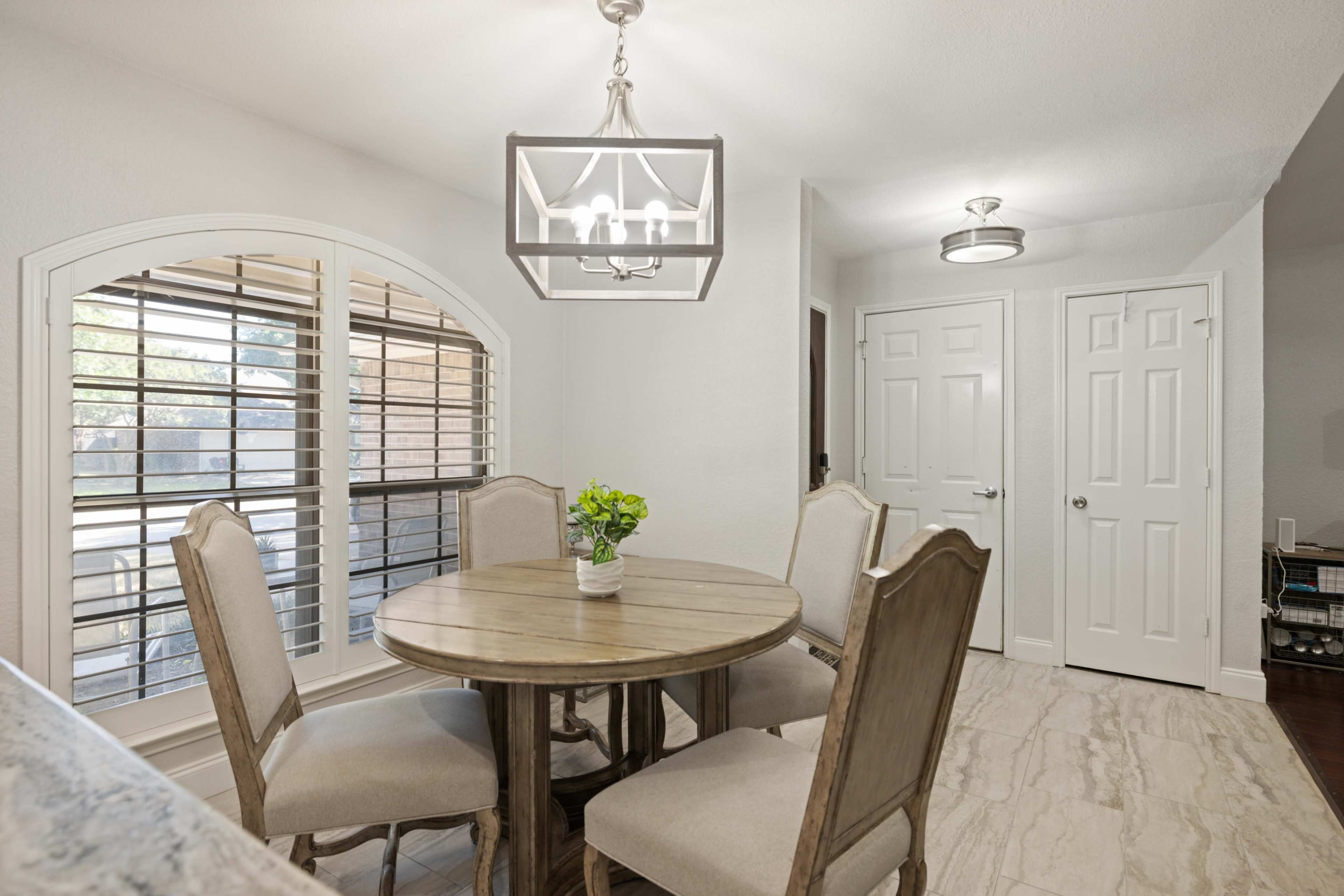 A dining area features a round wooden table surrounded by four upholstered chairs, with a light fixture above and shutters on the windows.