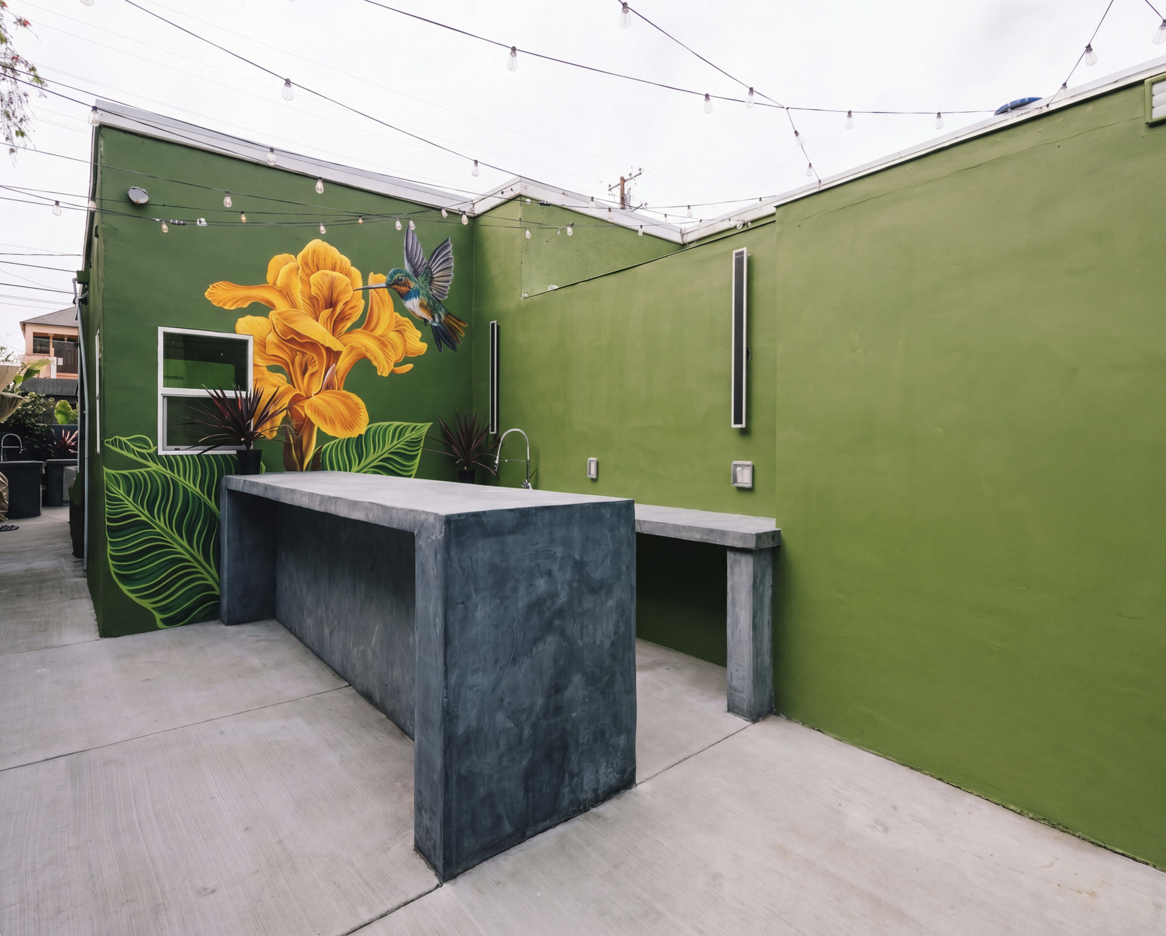 The image shows an outdoor space with a green wall featuring a large flower mural, alongside a concrete bar and modern lighting.