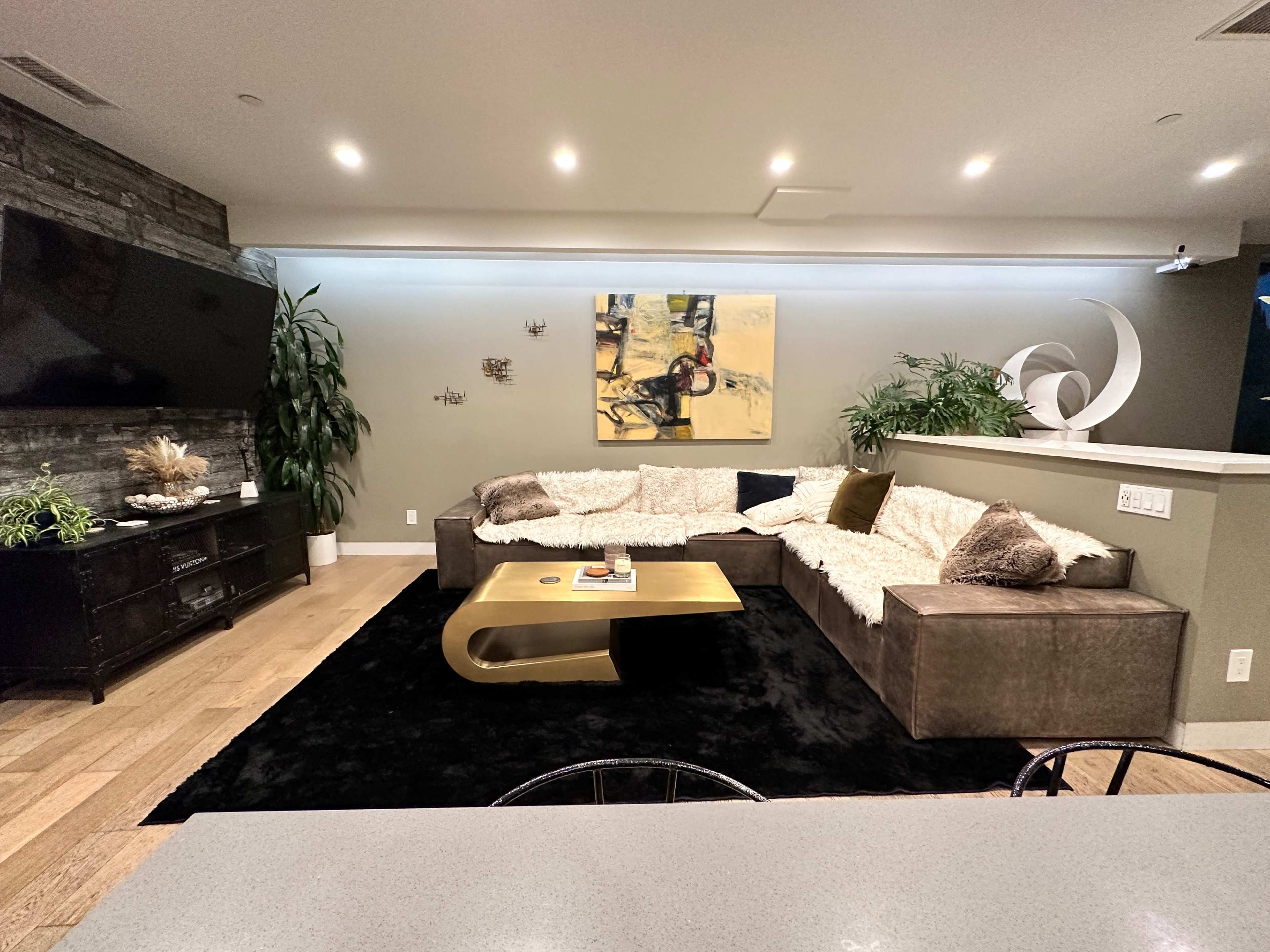 The image shows a modern living room with a large sectional sofa covered in a light textile, a coffee table, and various plants against a backdrop of a flat-screen TV and art on the walls.