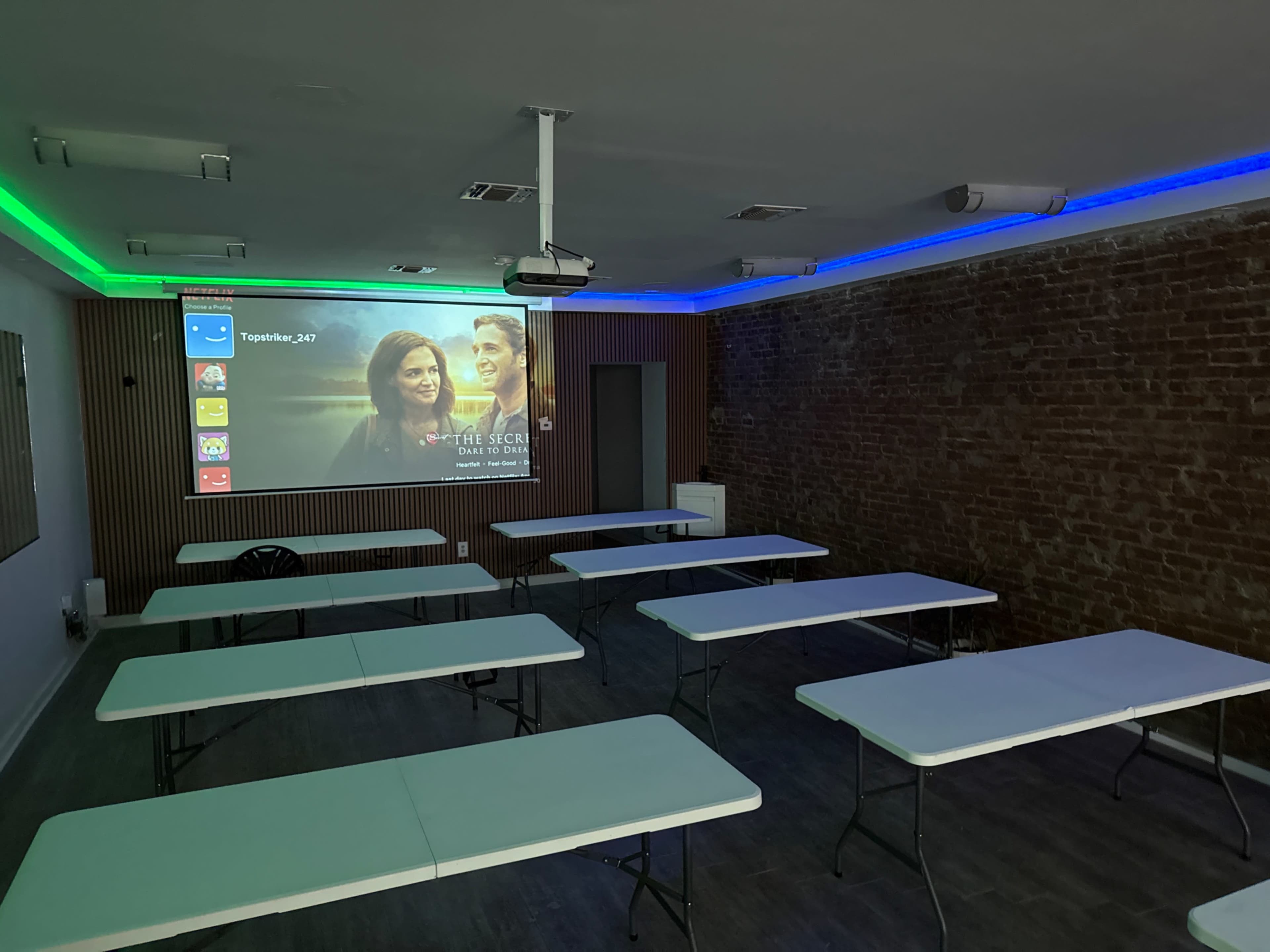 The image shows a classroom or meeting space featuring several empty folding tables arranged in rows, a projector screen displaying a movie poster, and LED light strips along the ceiling.