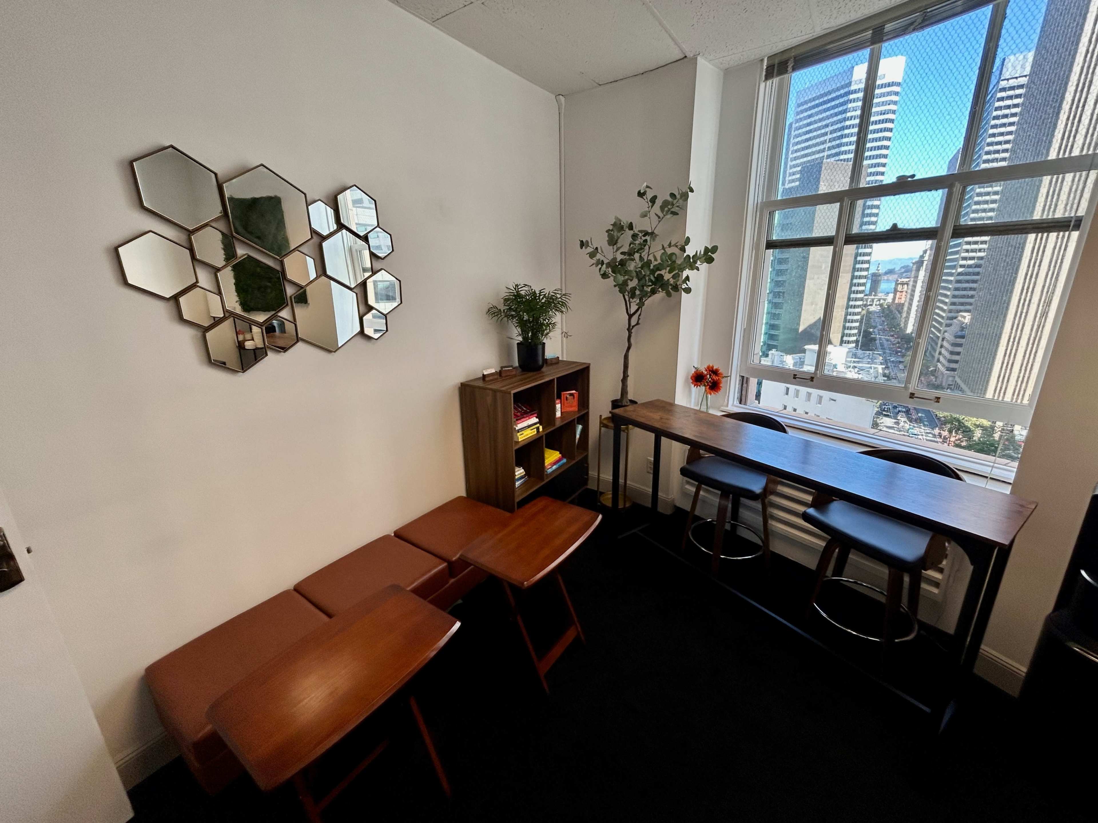 A small office space with a couple of wooden tables, a bookshelf, a vase with flowers, and a large window overlooking a city skyline.