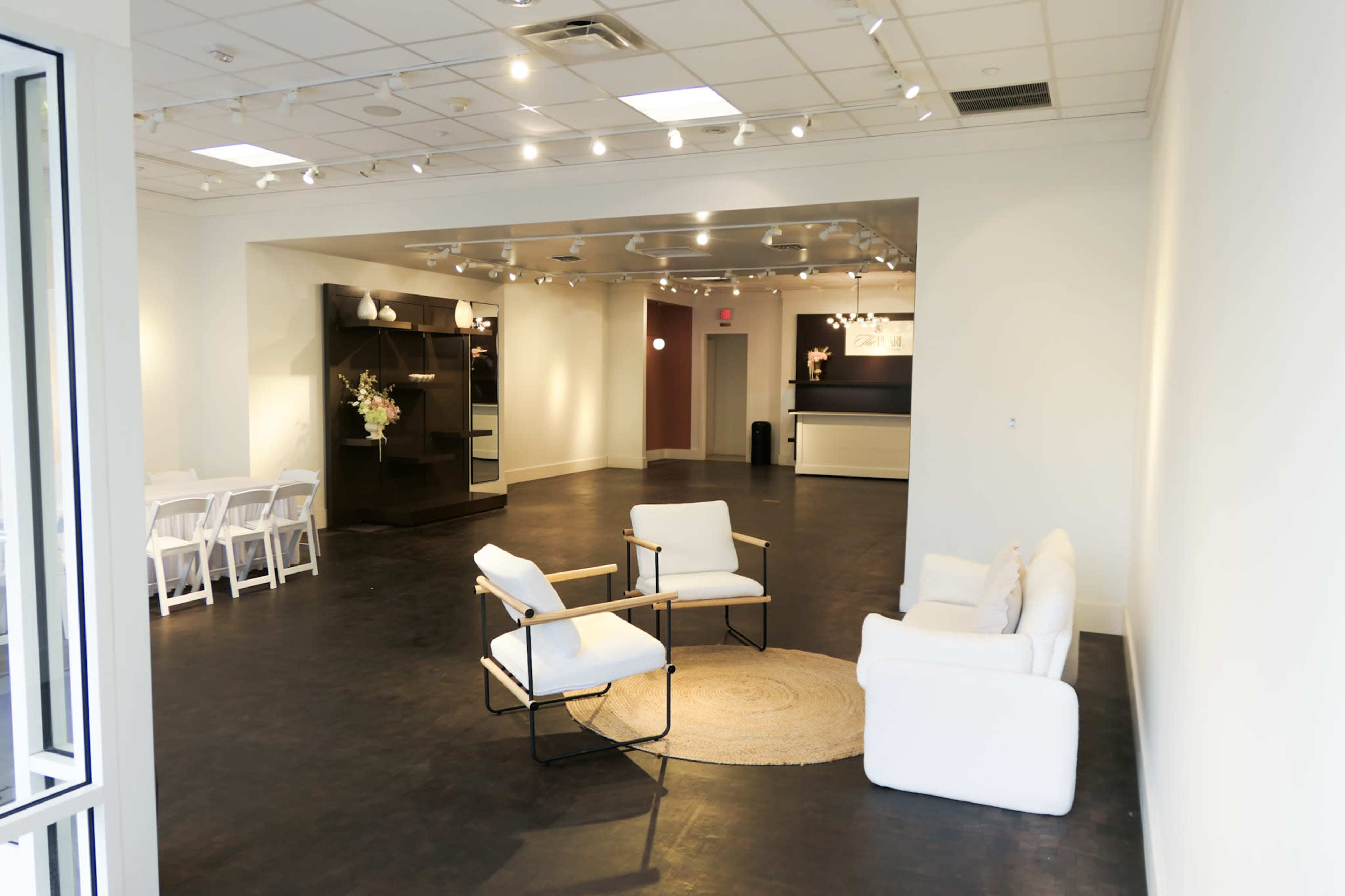 The image shows a spacious, well-lit interior with white walls, a seating area consisting of two chairs and a sofa, and a section featuring tables and decorative elements.
