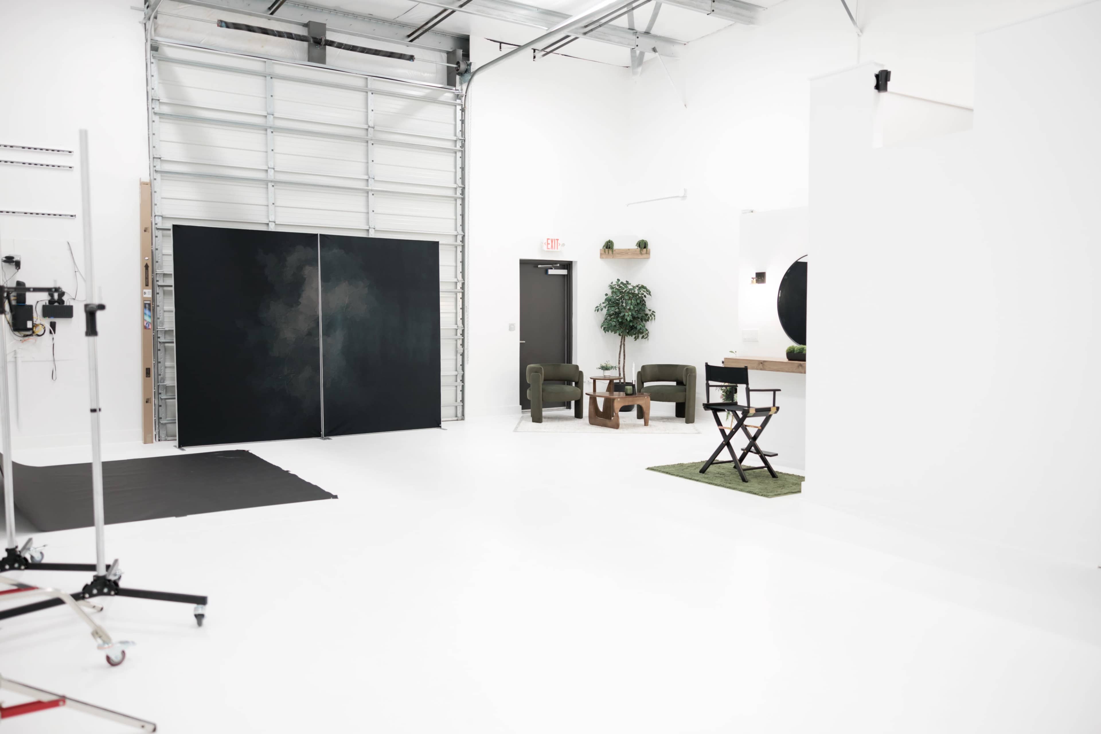 The image shows a spacious, clean studio with white walls, a concrete floor, and minimal furniture, including two green chairs, a small table, and a decorative plant.