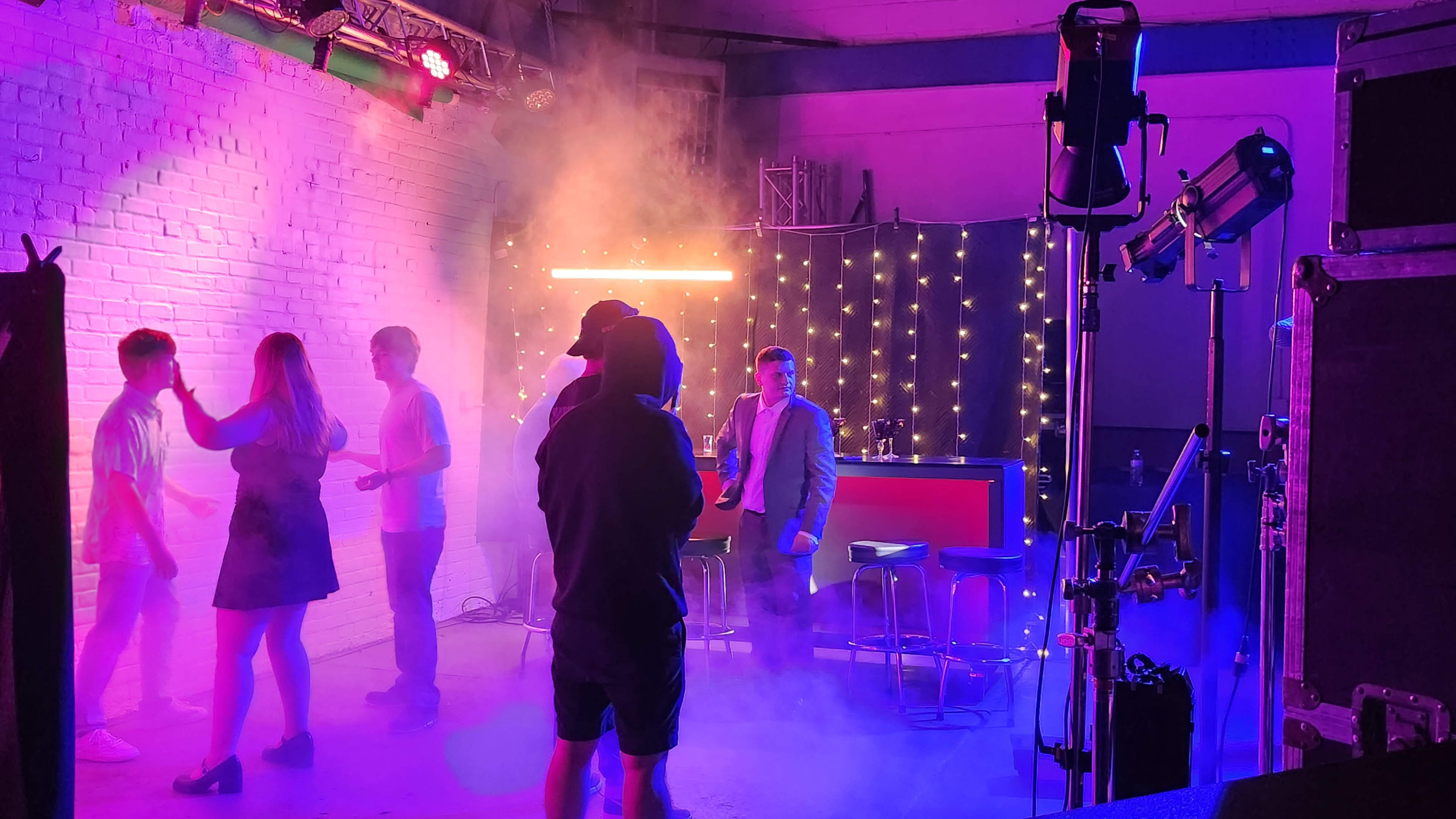 A group of people are interacting on a stage set illuminated by colorful lights and surrounded by smoke.
