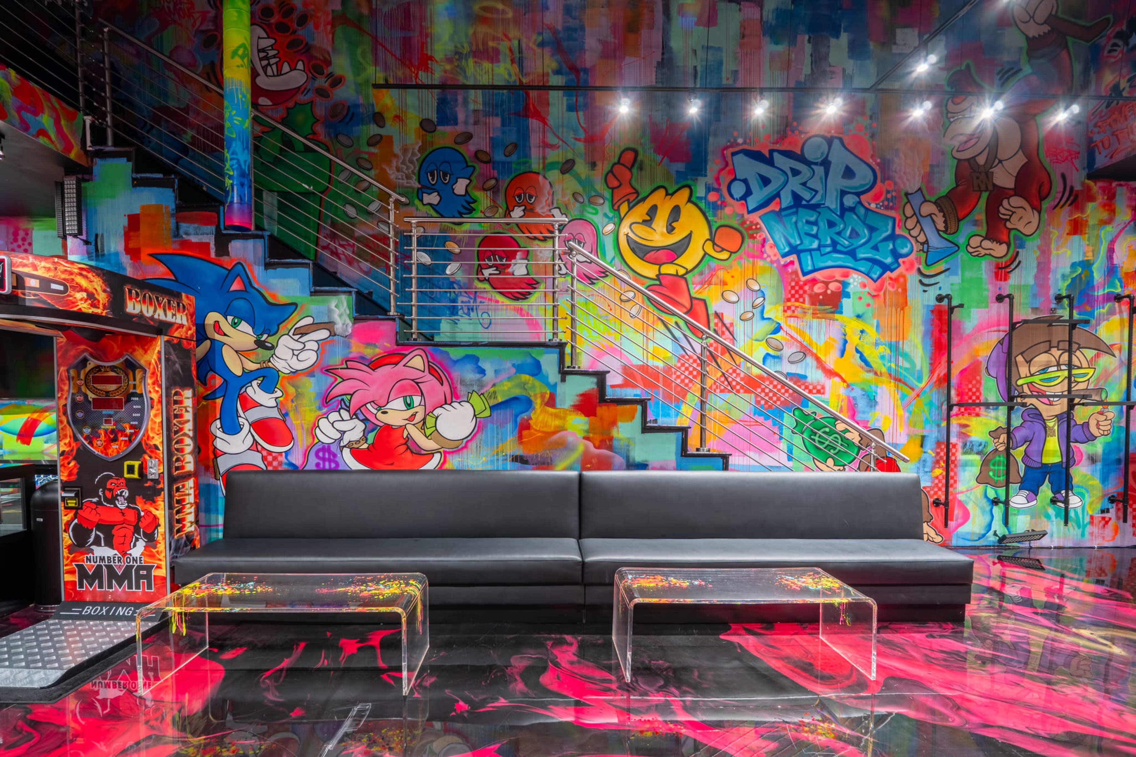 The image shows a vibrant gaming room featuring colorful graffiti art on the walls, a black couch, and clear plastic tables.