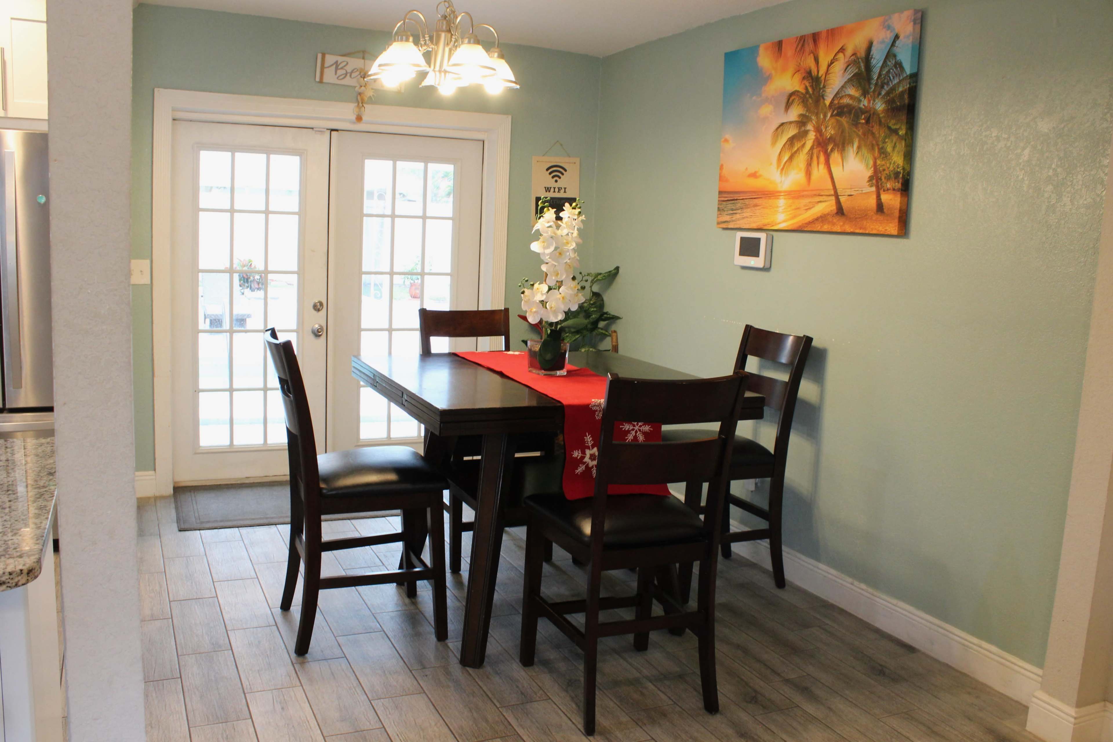 A dining area with a rectangular table set for four, a red table runner, and a view of a bright tropical-themed painting on the wall beside a pair of double glass doors.