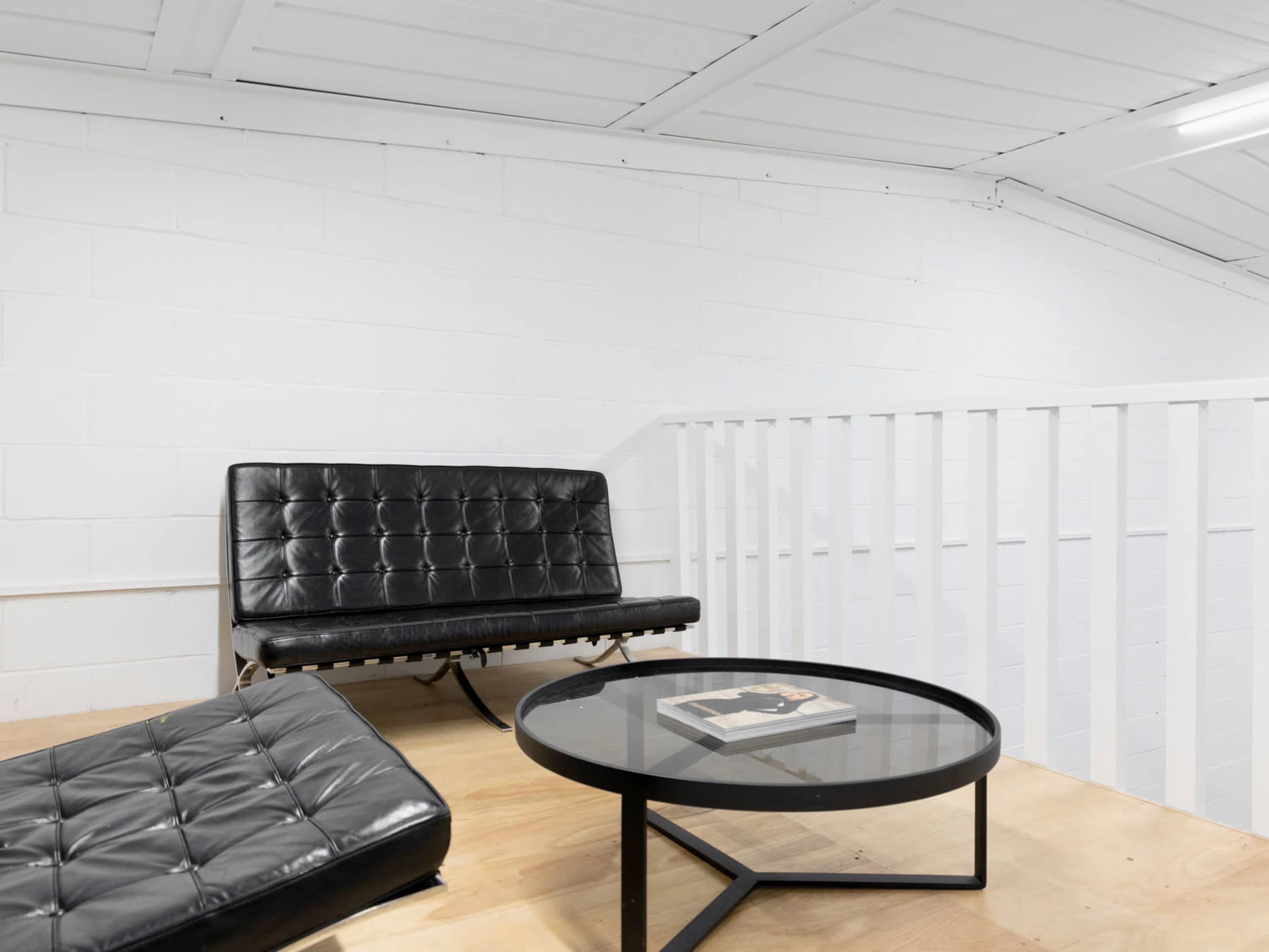 A minimalist living space with a black leather sofa, a low glass coffee table, and a wooden floor beneath white walls and ceiling.