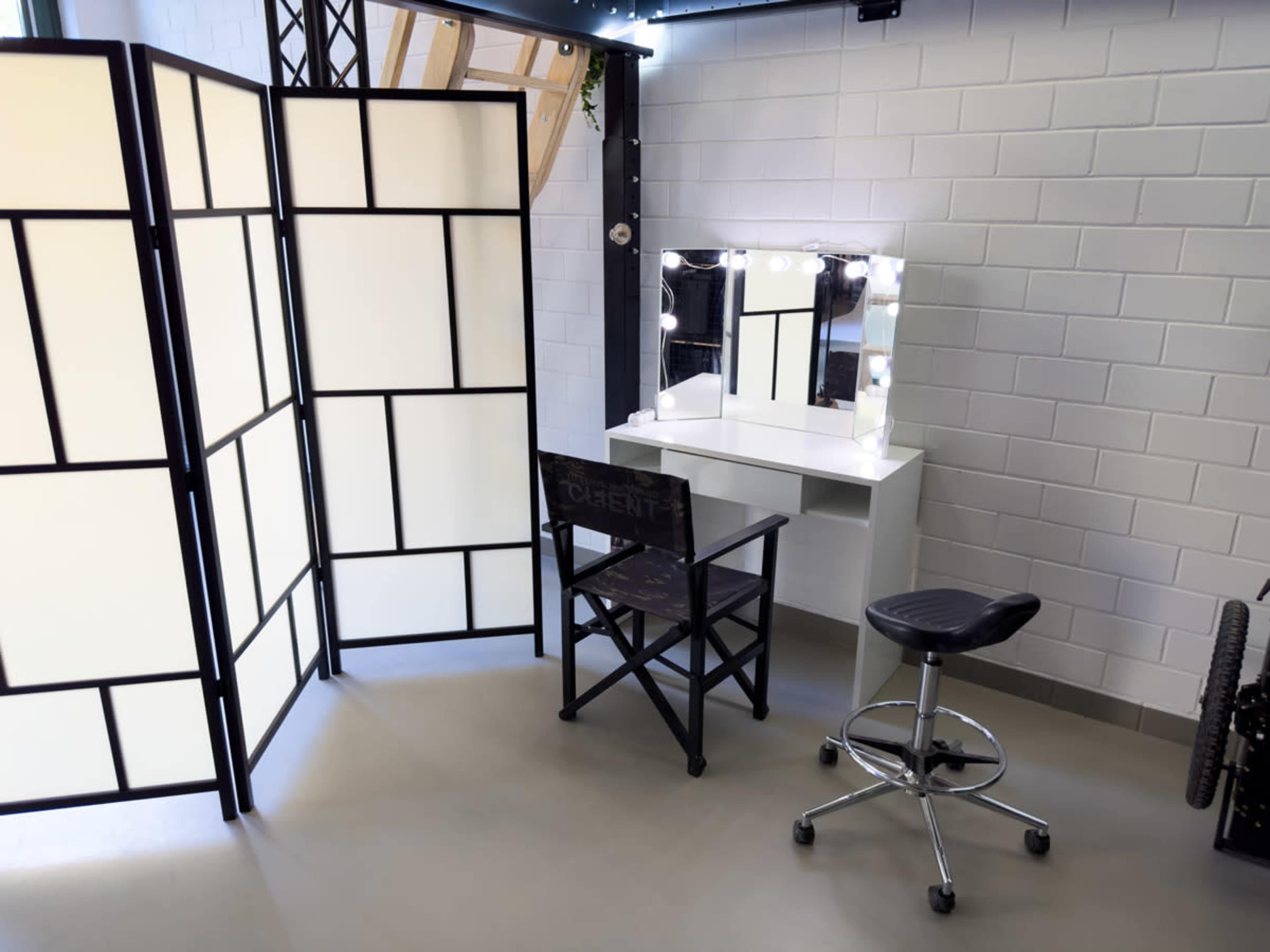 A makeup station with a illuminated mirror, a director's chair, and a rolling stool surrounded by a folding screen.