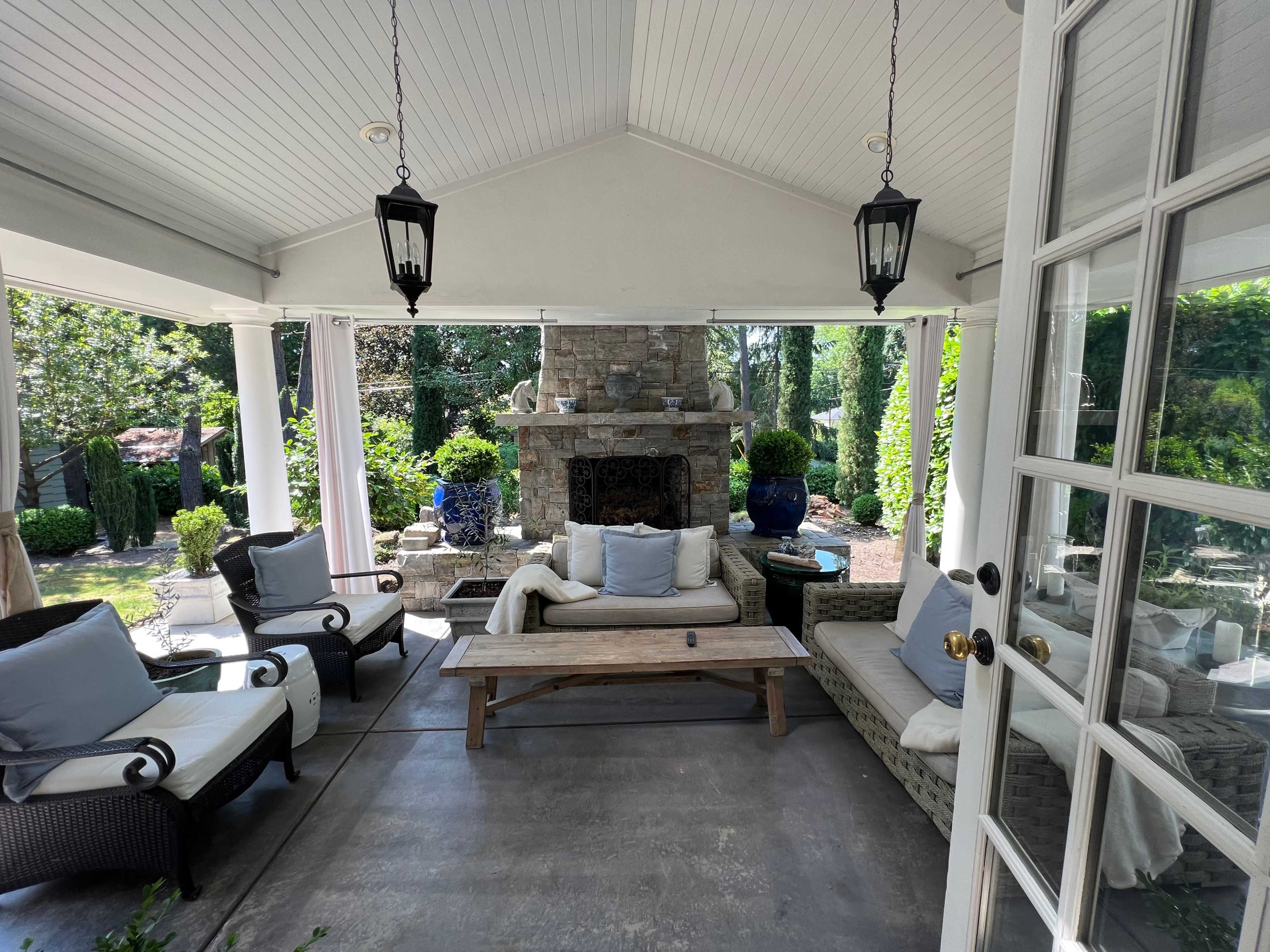 A covered outdoor sitting area with a stone fireplace, wicker furniture, and lush greenery in the background.