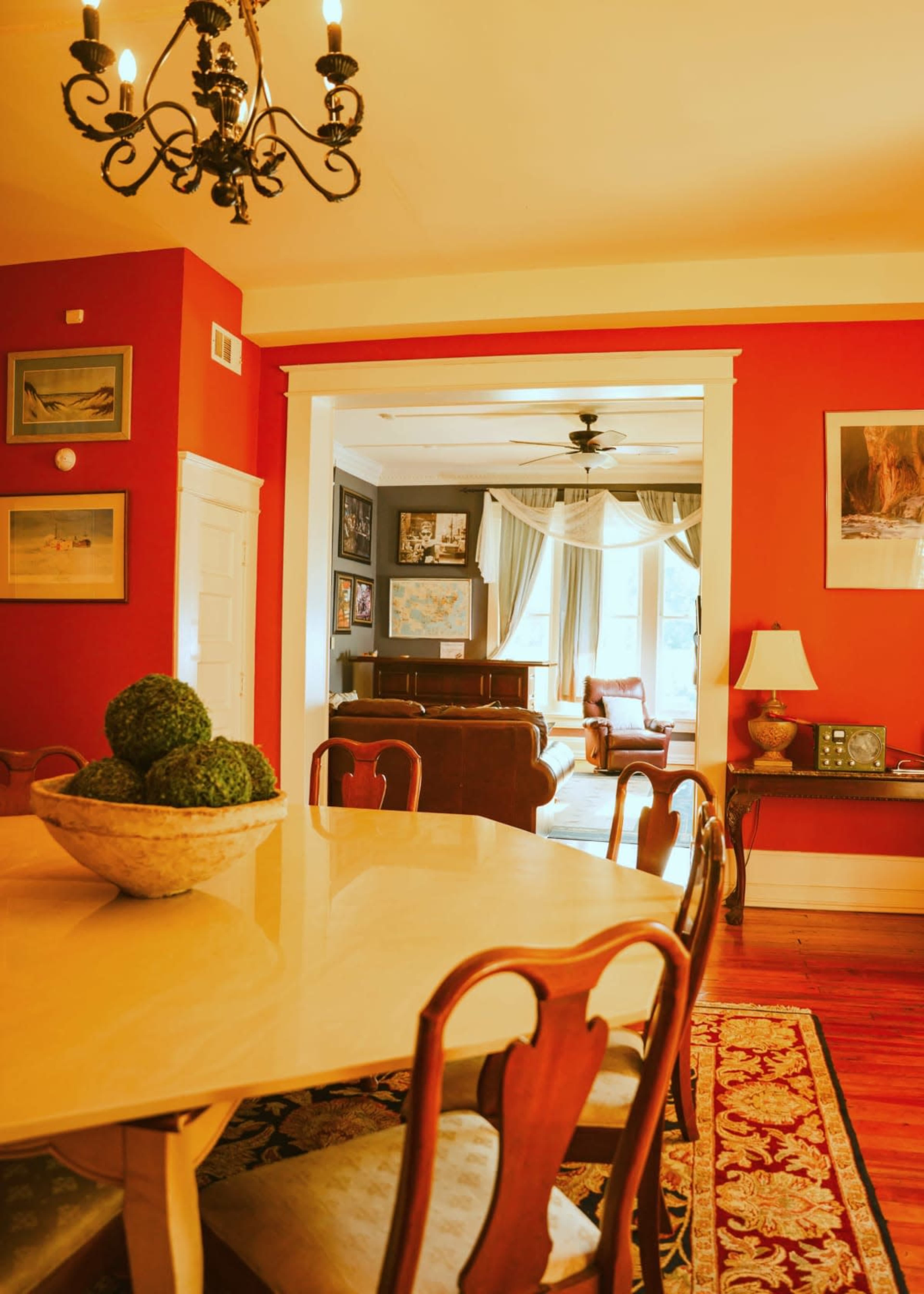 The image shows a dining room with a round table and a chandelier, leading into a cozy living room adorned with framed pictures and a large window.