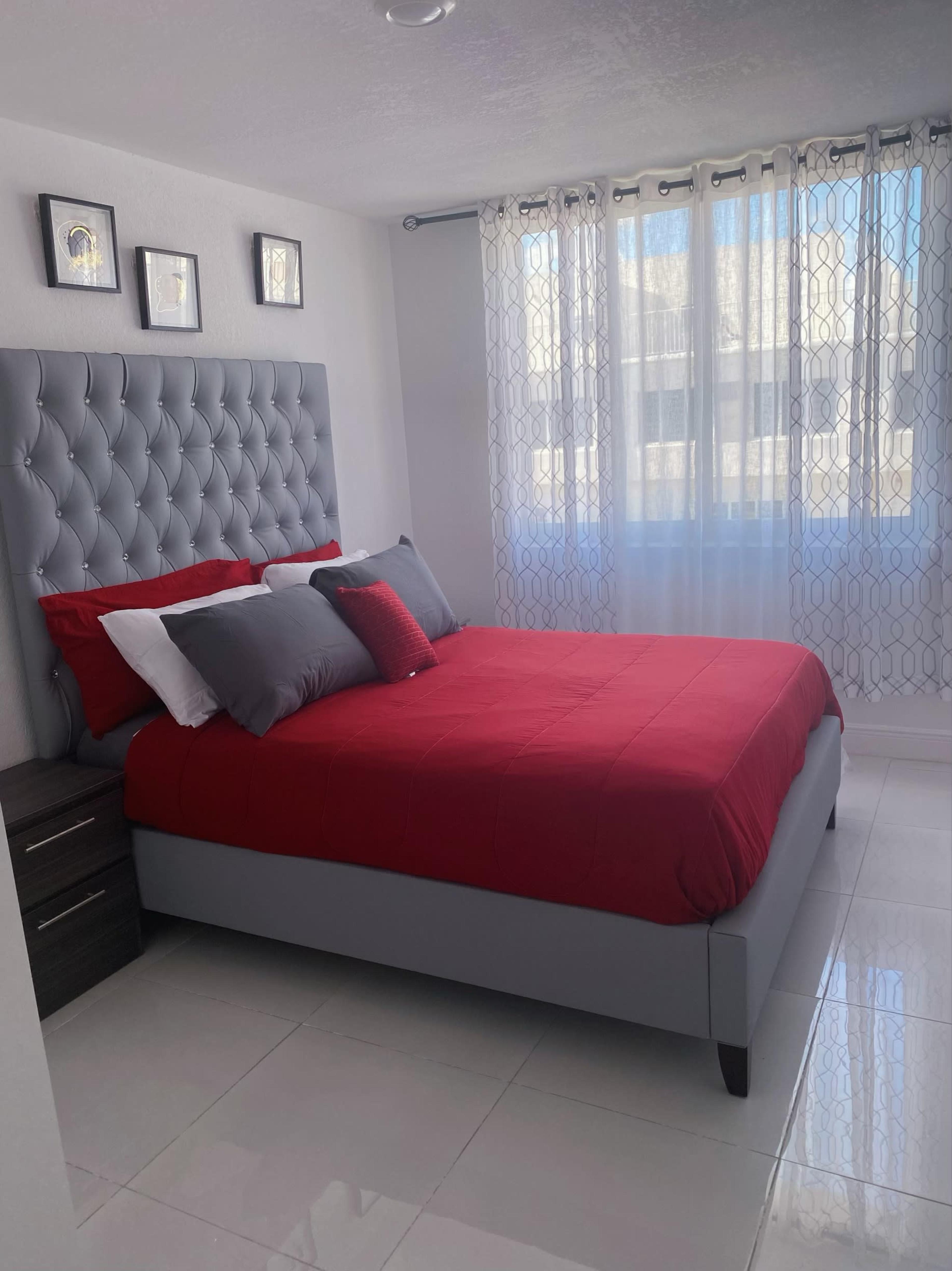 A neatly arranged bedroom featuring a gray upholstered bed with red and gray bedding, white walls, and large windows adorned with sheer curtains.