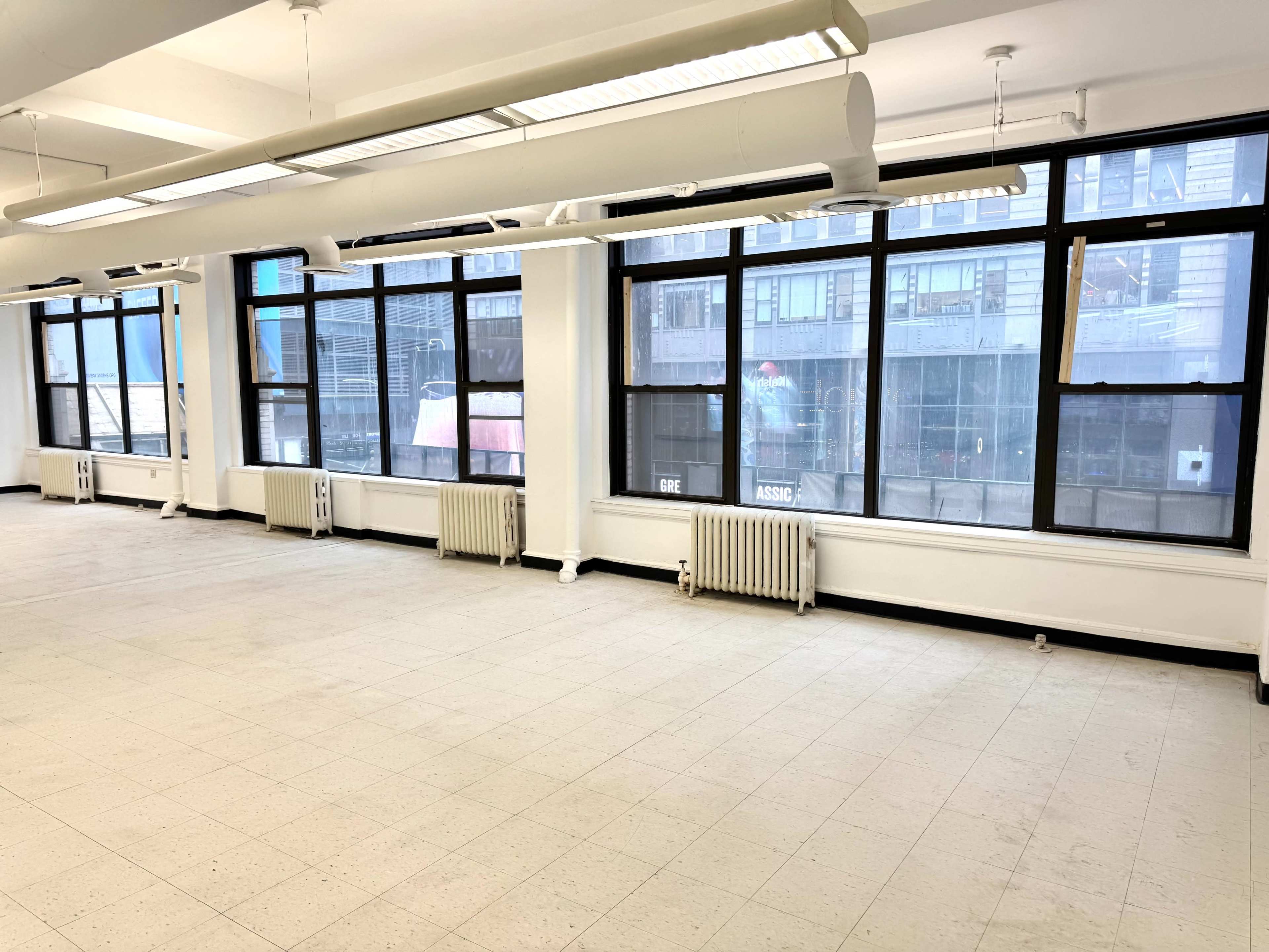 The image shows a large, empty room with multiple windows and radiator heating along the walls.