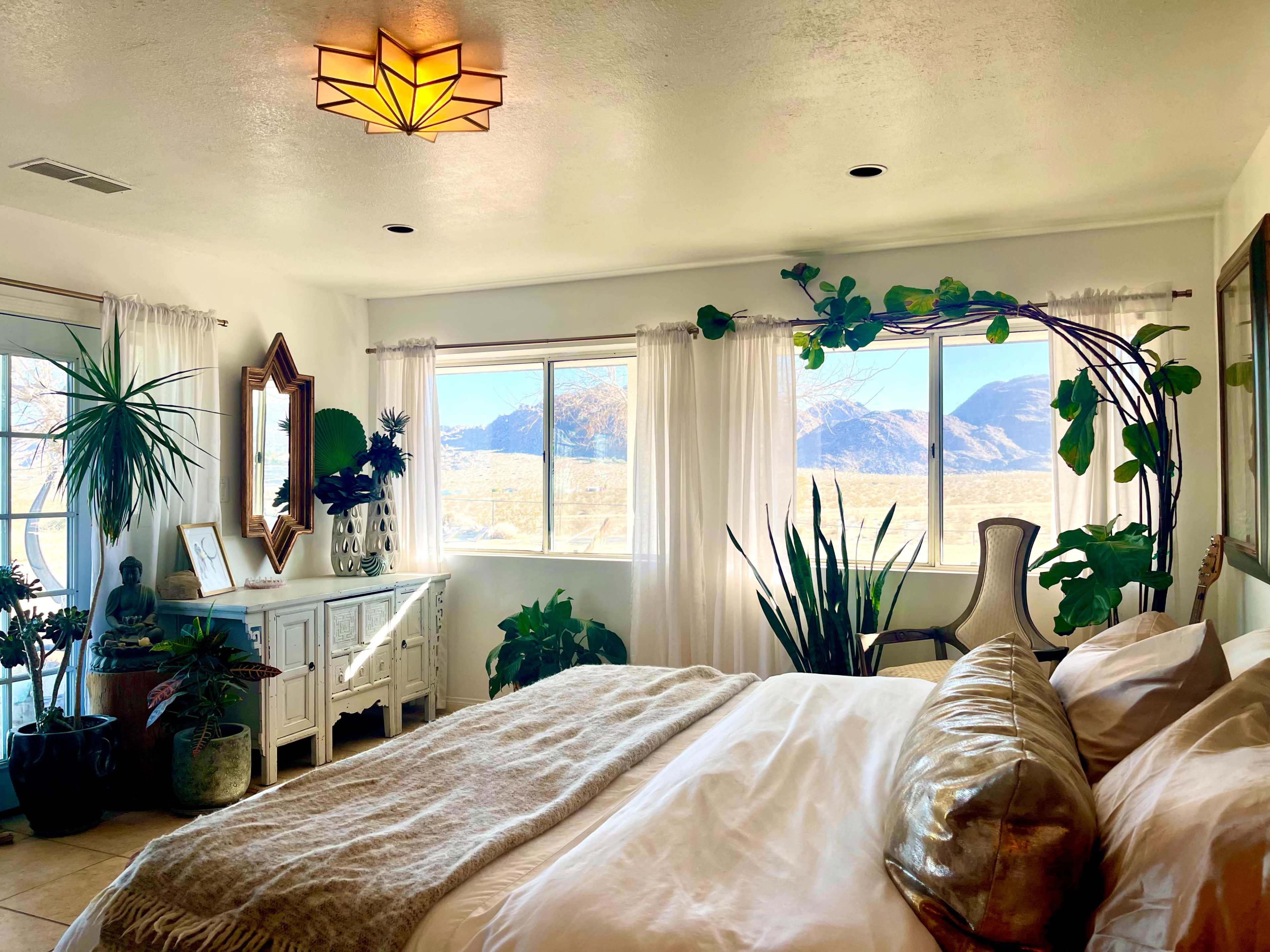 A bright bedroom features large windows showcasing a mountain view, adorned with various plants and minimalist decor.