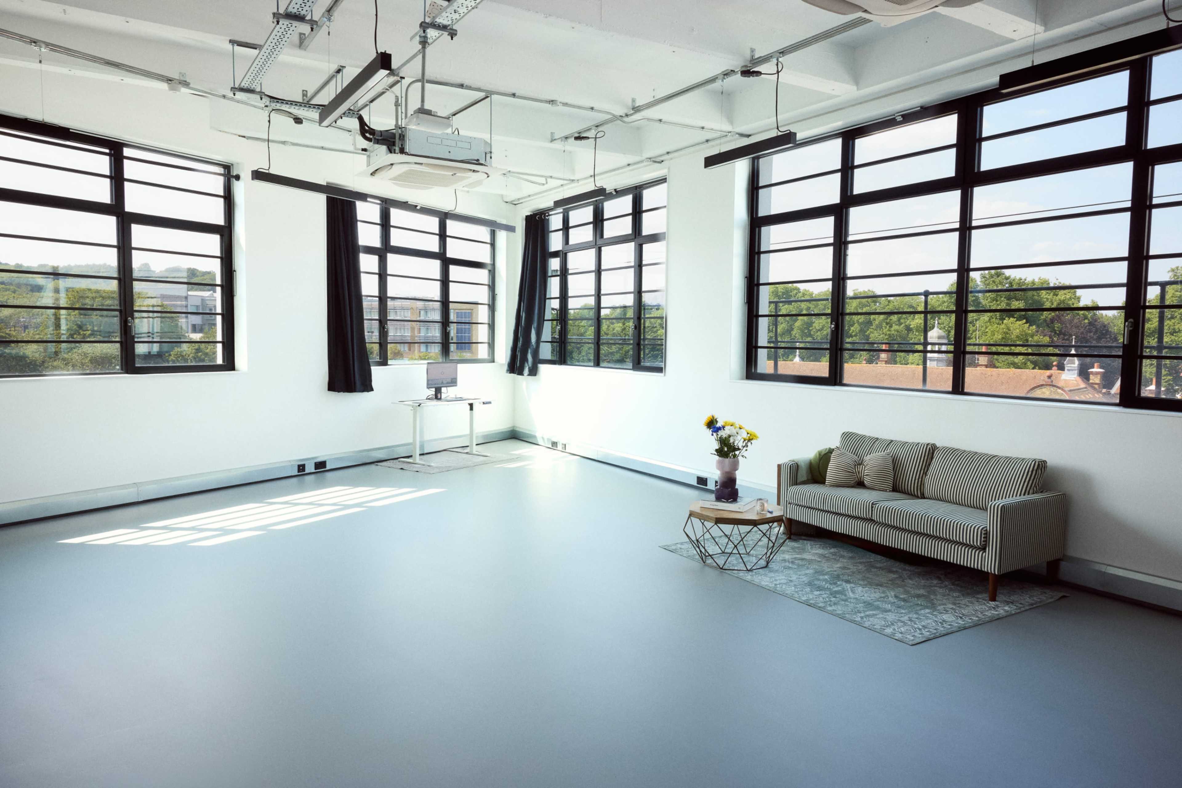The image shows a bright, spacious room with large windows, a couch, a small table with flowers, and a desk against the wall.