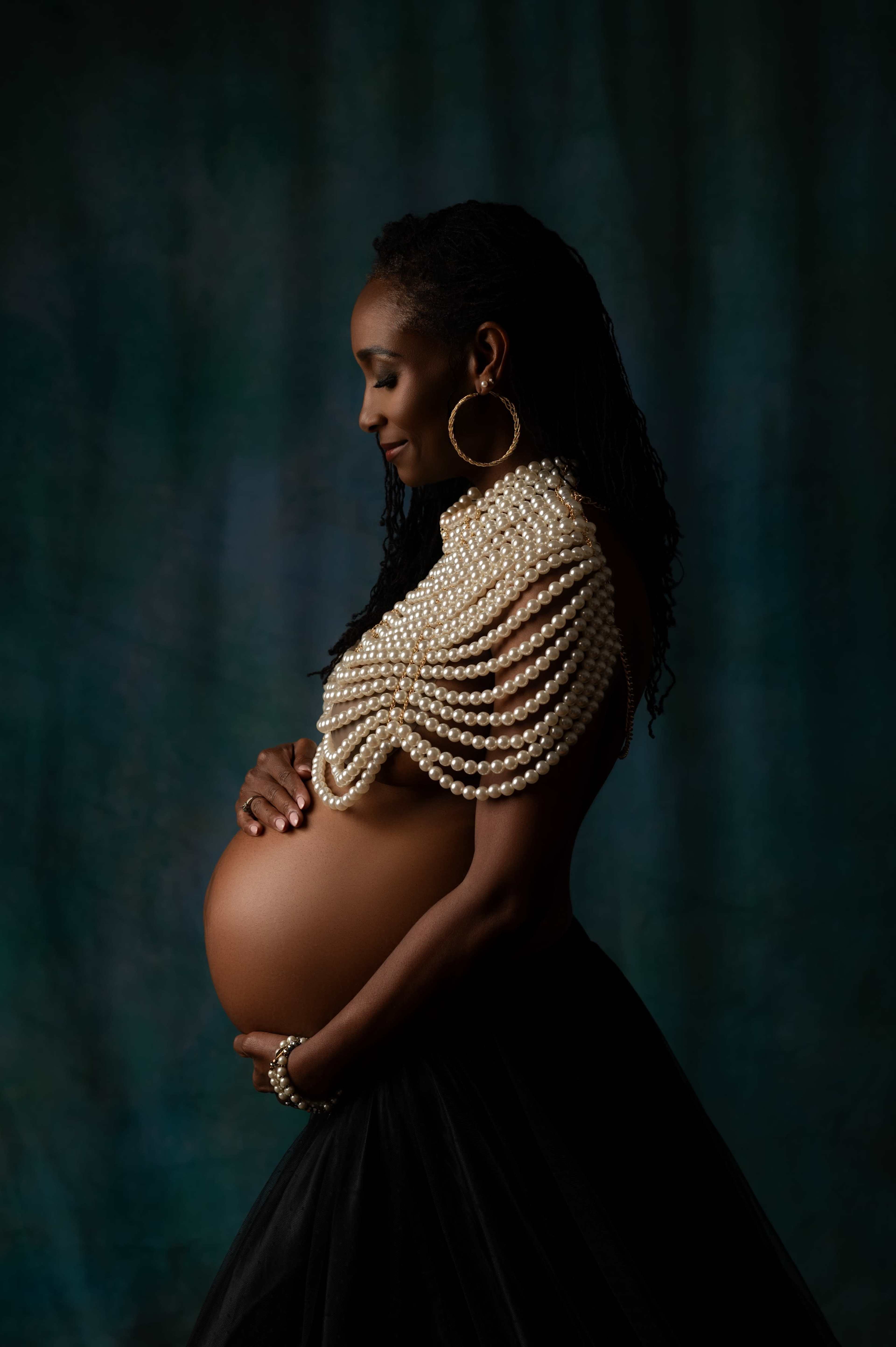 A pregnant woman in profile stands gracefully, adorned with a cascade of pearls and wearing a sheer black skirt against a textured blue-green backdrop.