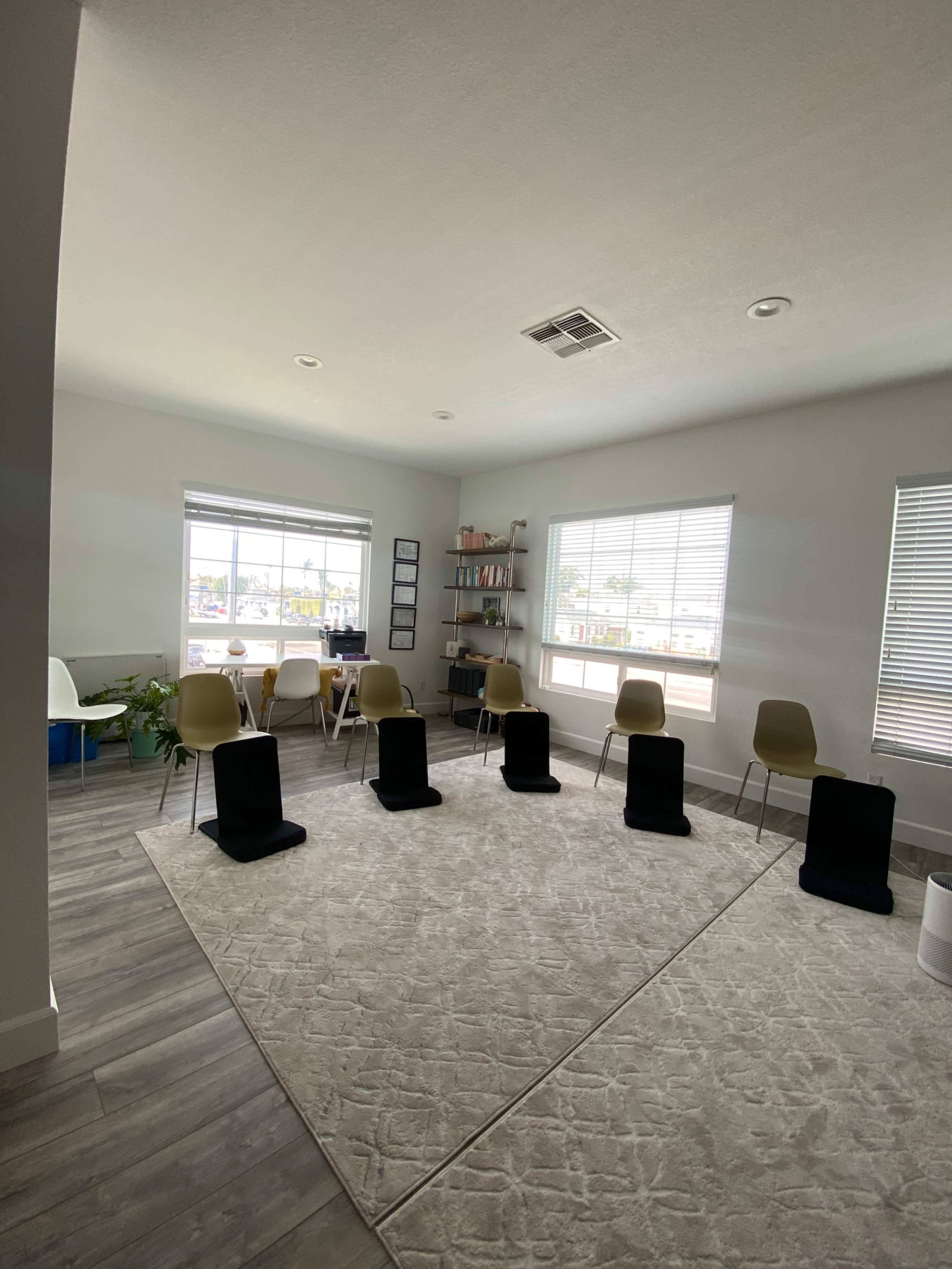 A modern room with a circular seating arrangement of black chairs on a light rug, surrounded by windows and a table with additional chairs.