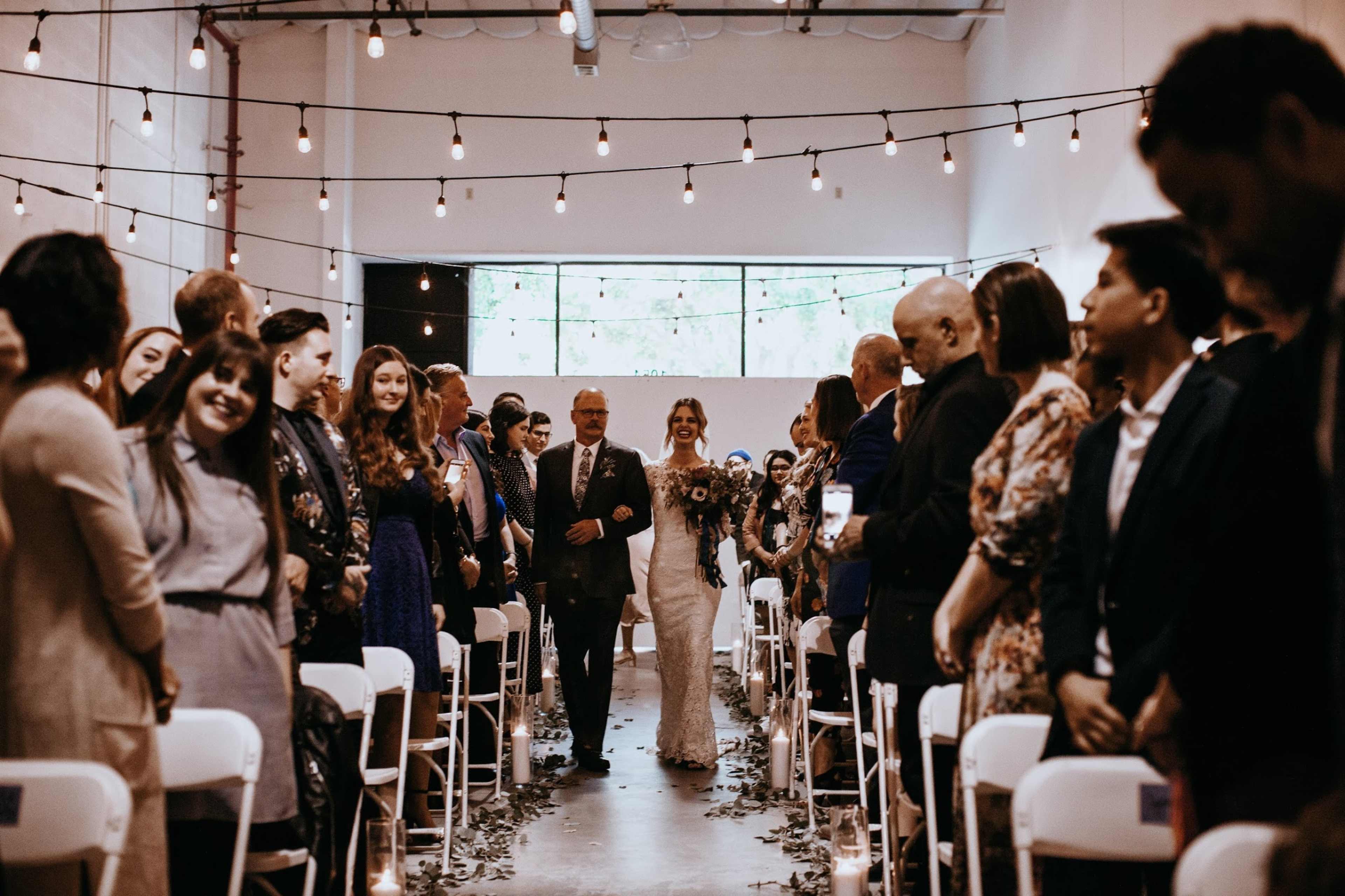 A bride walks down the aisle accompanied by her father, with guests seated on either side under string lights in a decorated venue.
