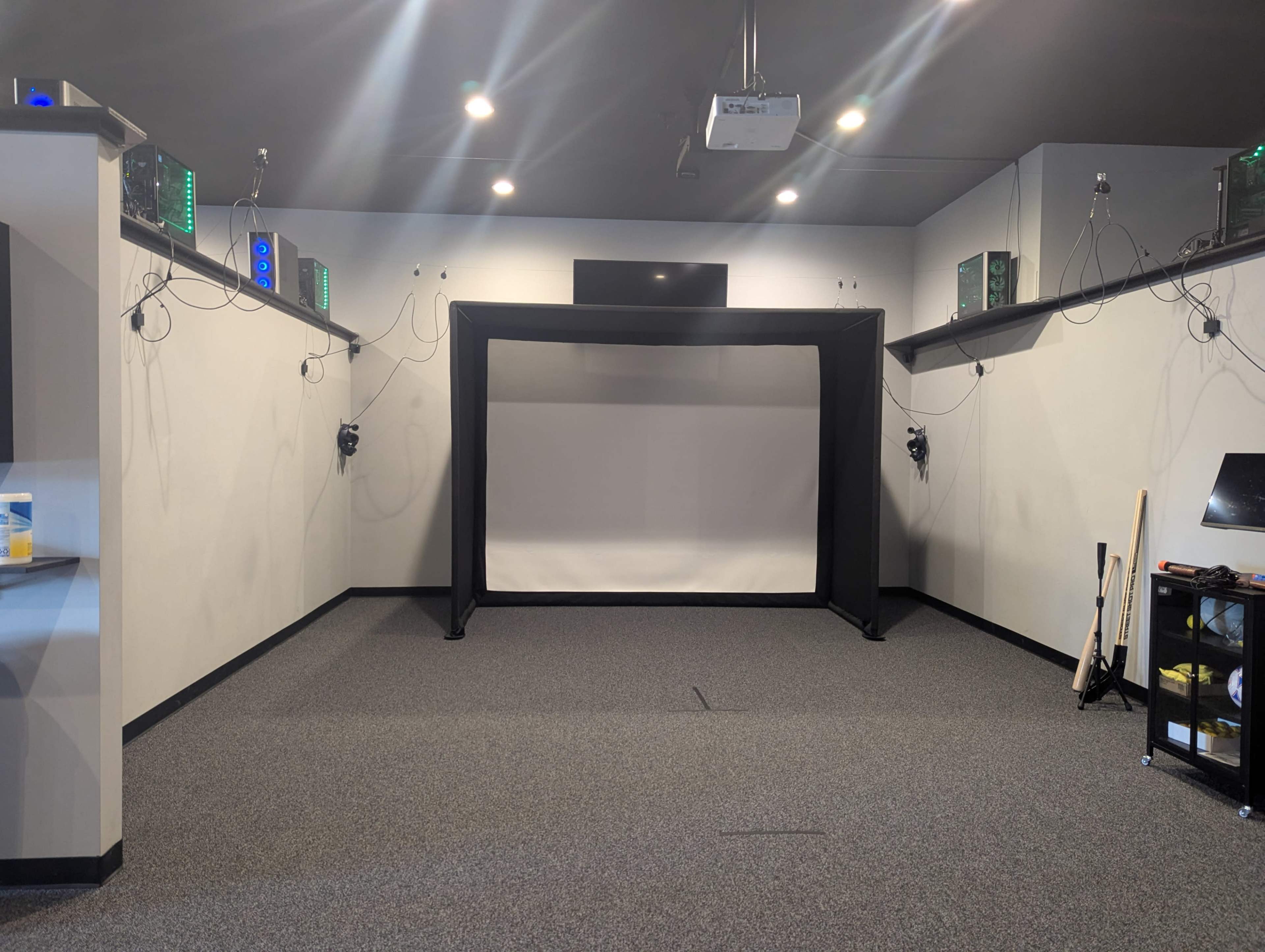 The image shows an empty room featuring a black projection screen, wall-mounted speakers, and a projector overhead, with a carpeted floor and minimal furnishings.