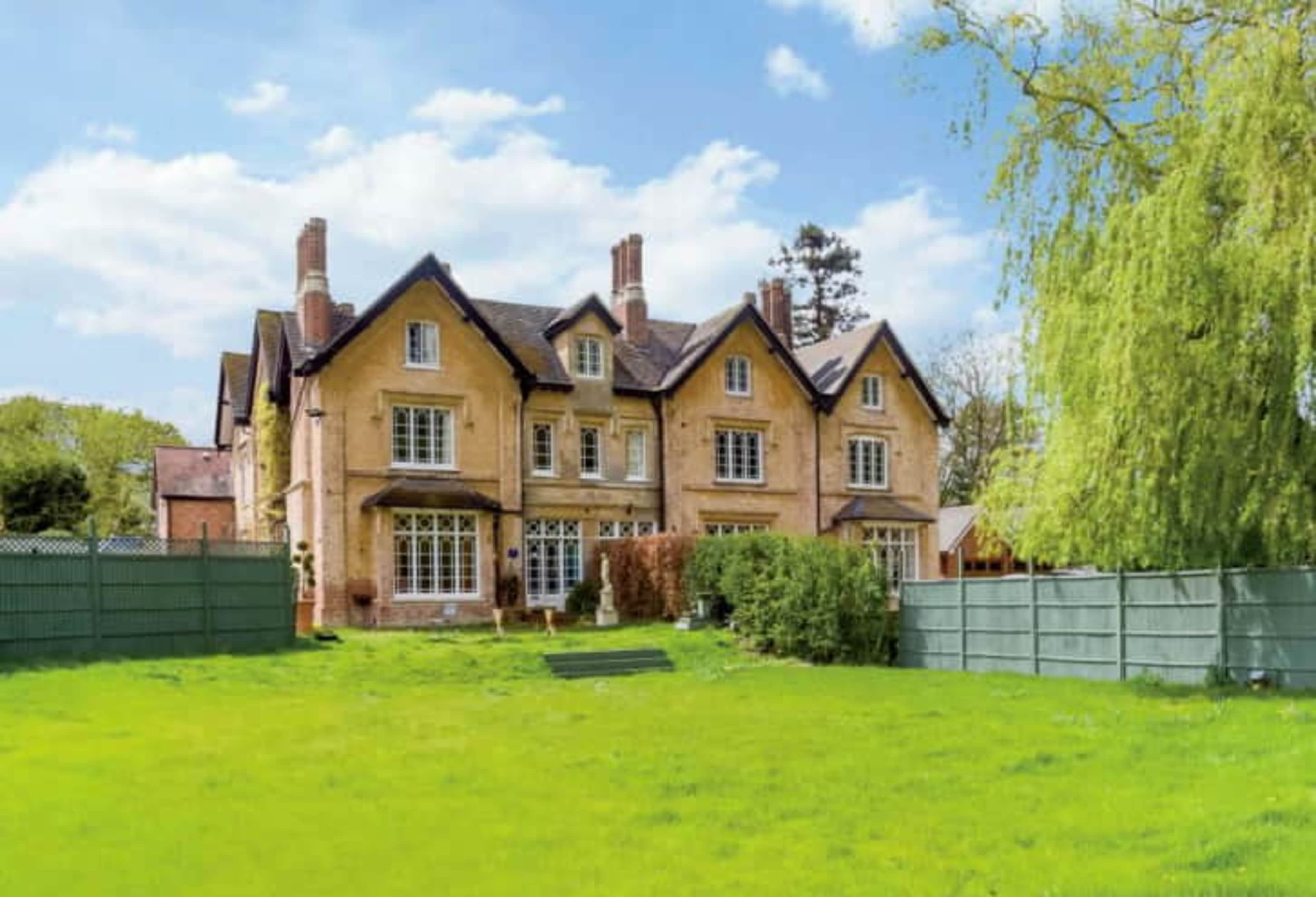 A large, three-story brick house is set on a green lawn surrounded by a wooden fence and trees.