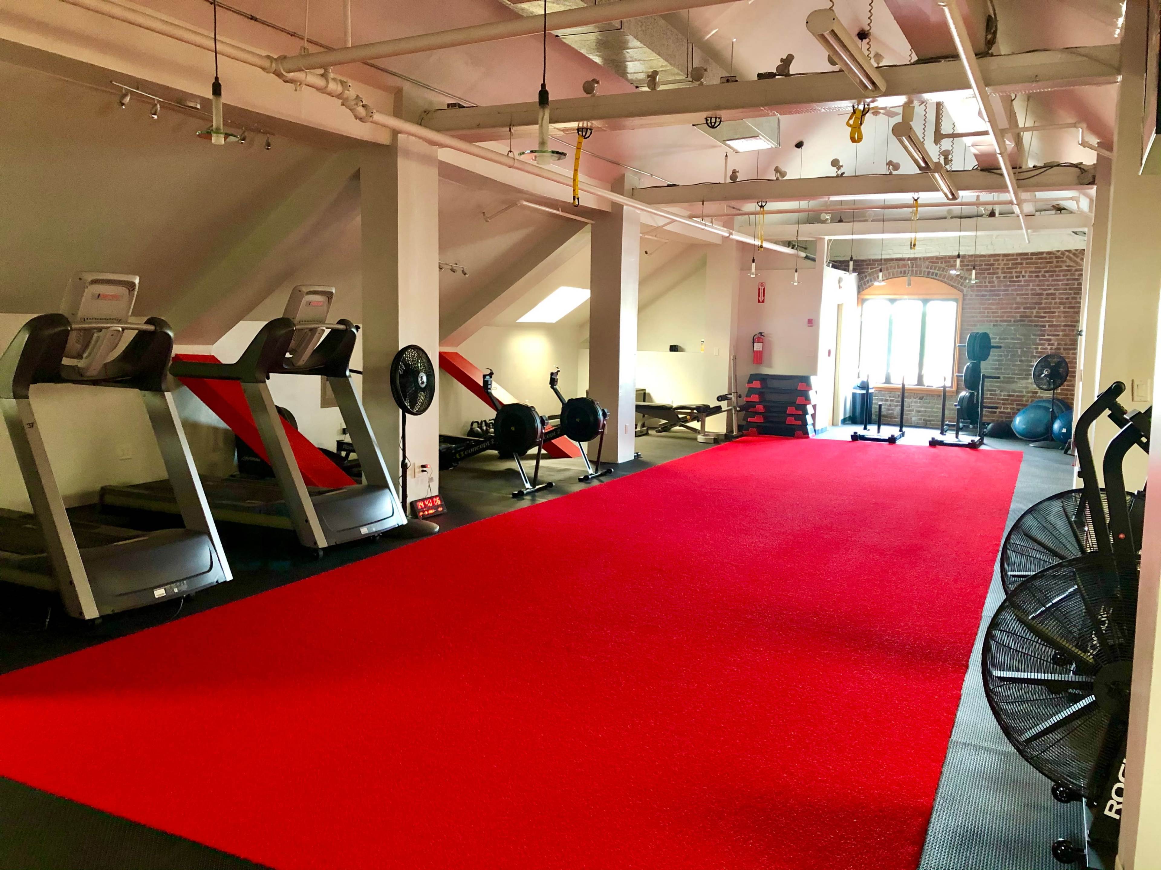 A modern gym interior featuring red carpeting, several treadmills, exercise bikes, and weightlifting equipment along with large windows allowing natural light.
