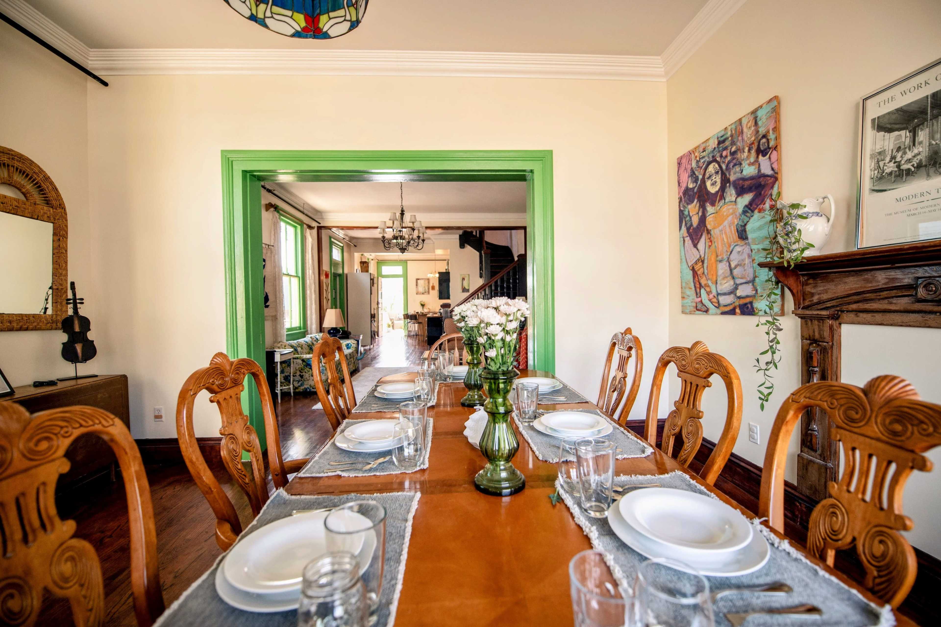 The image shows a spacious dining room with a long wooden table set for a meal, featuring finely arranged plates and glasses, with an open doorway leading to a well-lit living area.