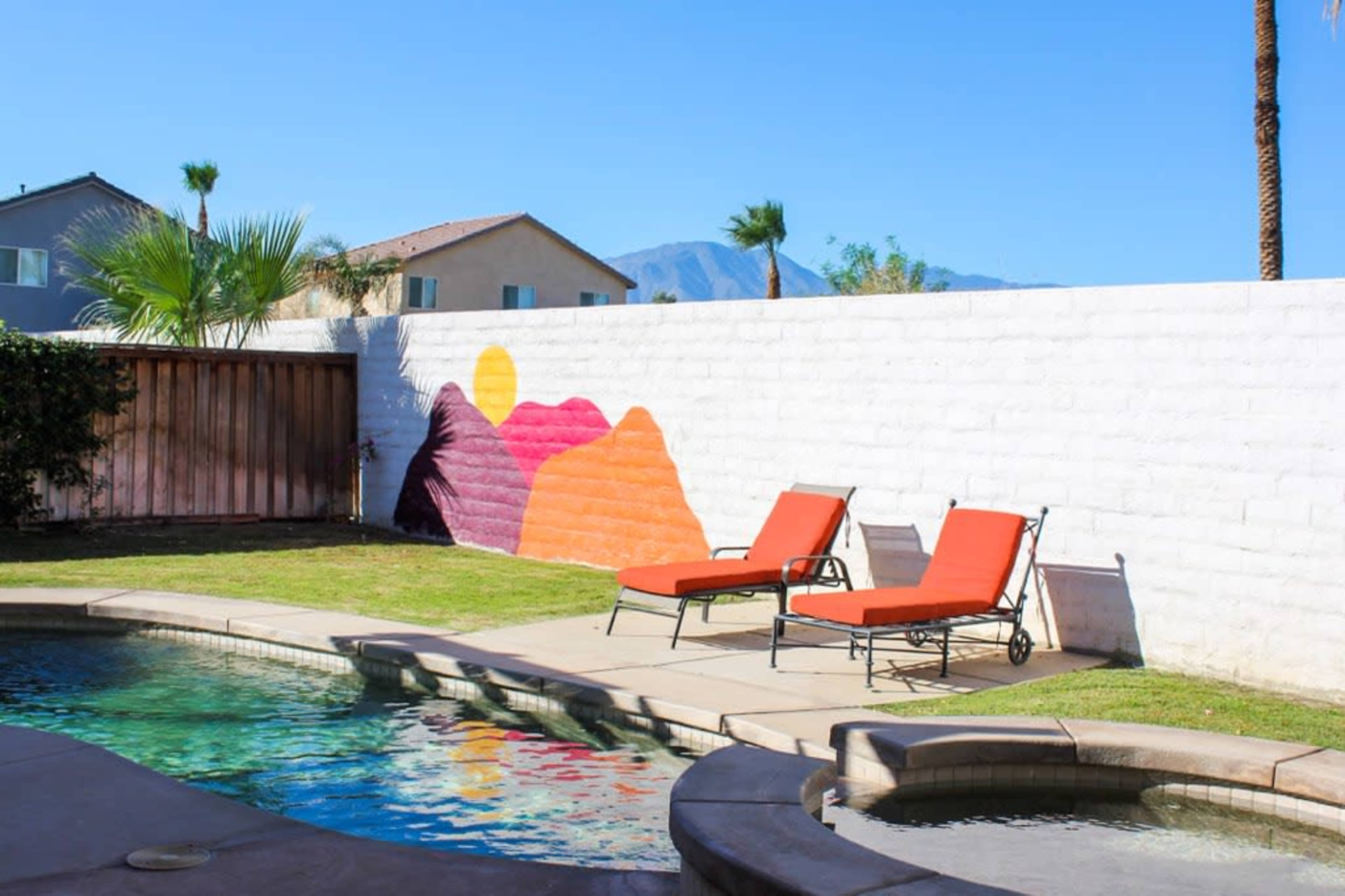 A swimming pool area with two orange lounge chairs beside a colorful mountain mural on a white wall.