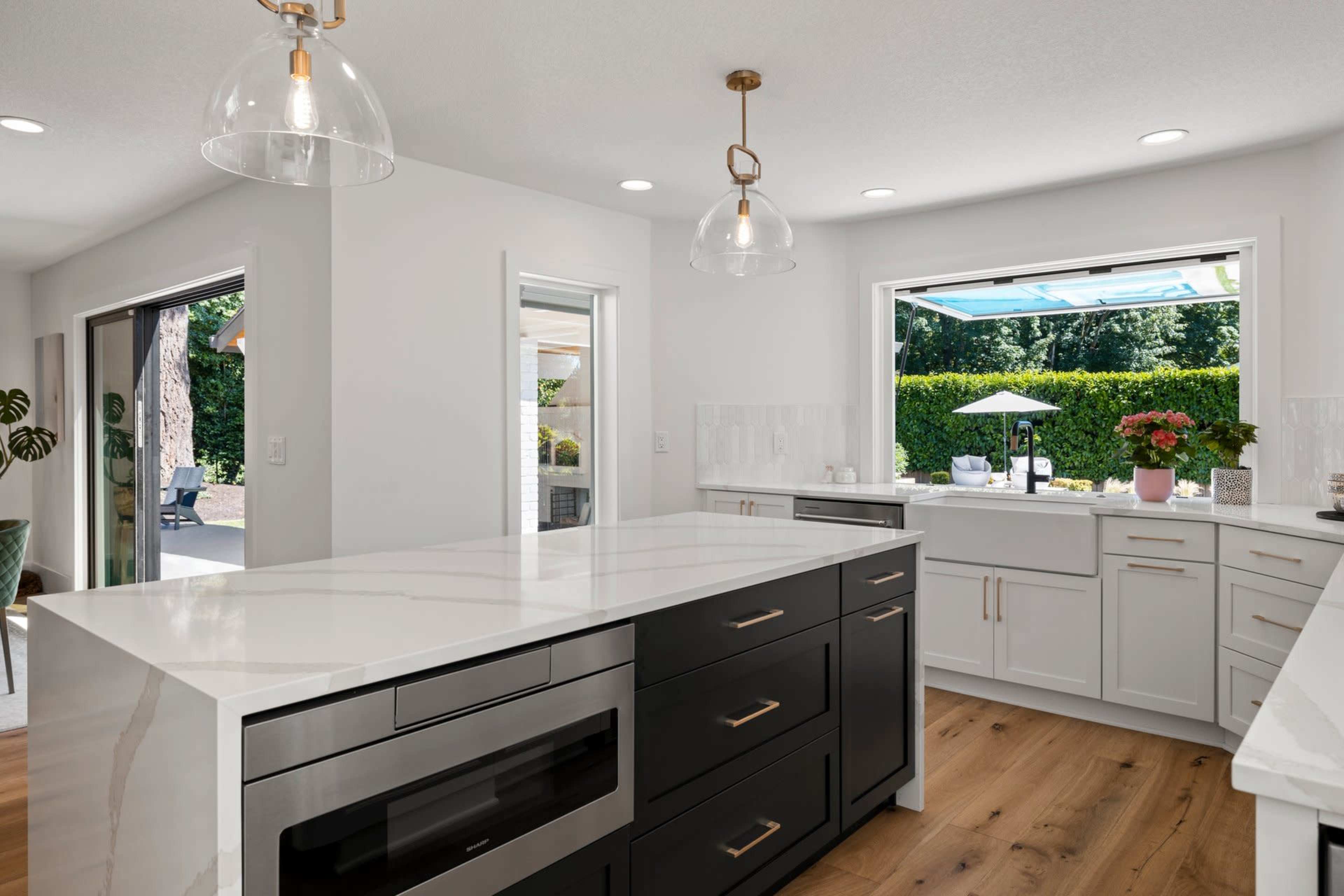 A modern kitchen with a large island, featuring a black countertop and stainless steel appliances, and a window that opens to an outdoor area.