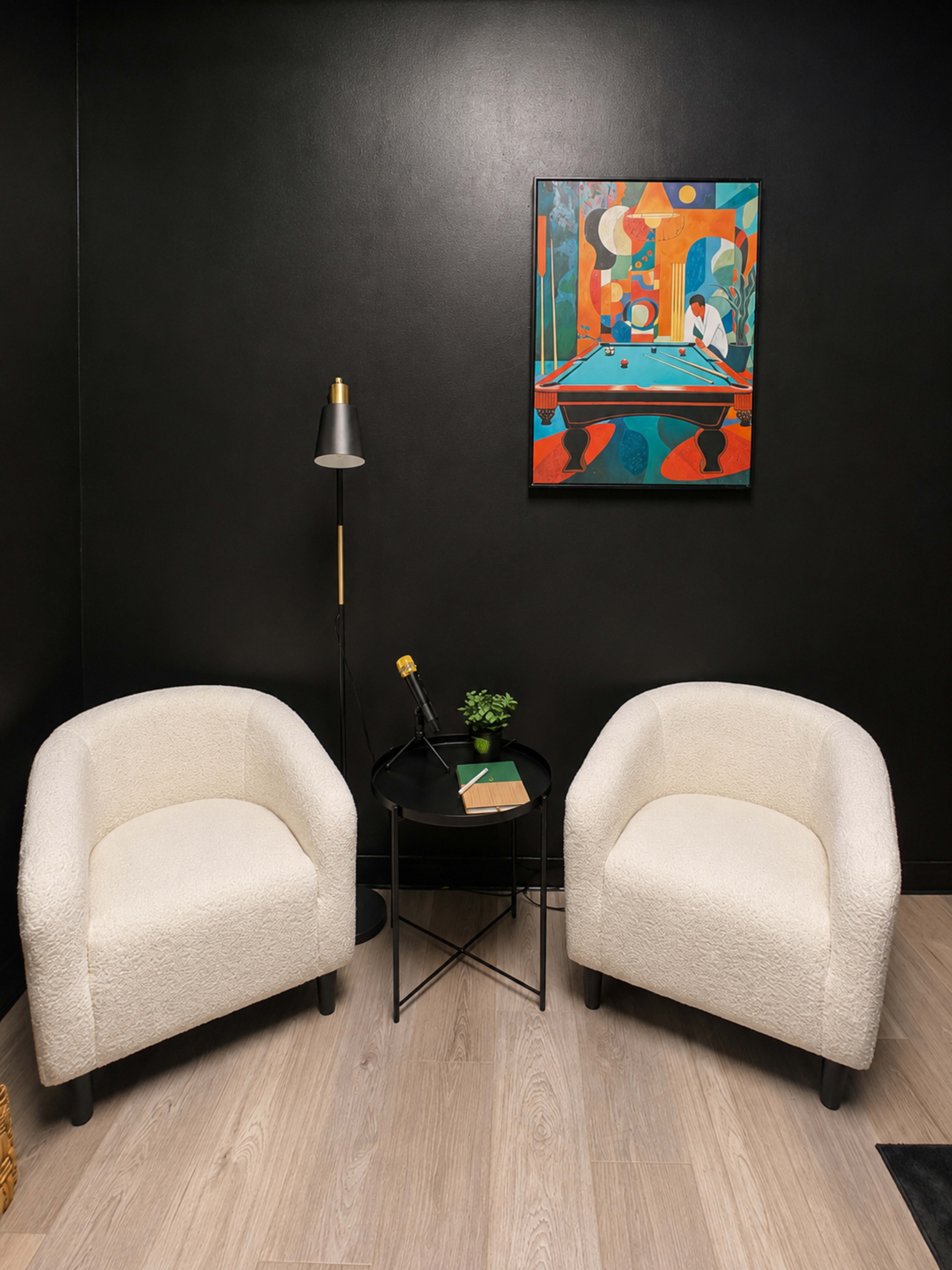The image shows a small, modern seating area with two white upholstered chairs, a round black side table, a floor lamp, and a colorful abstract painting on a black wall.