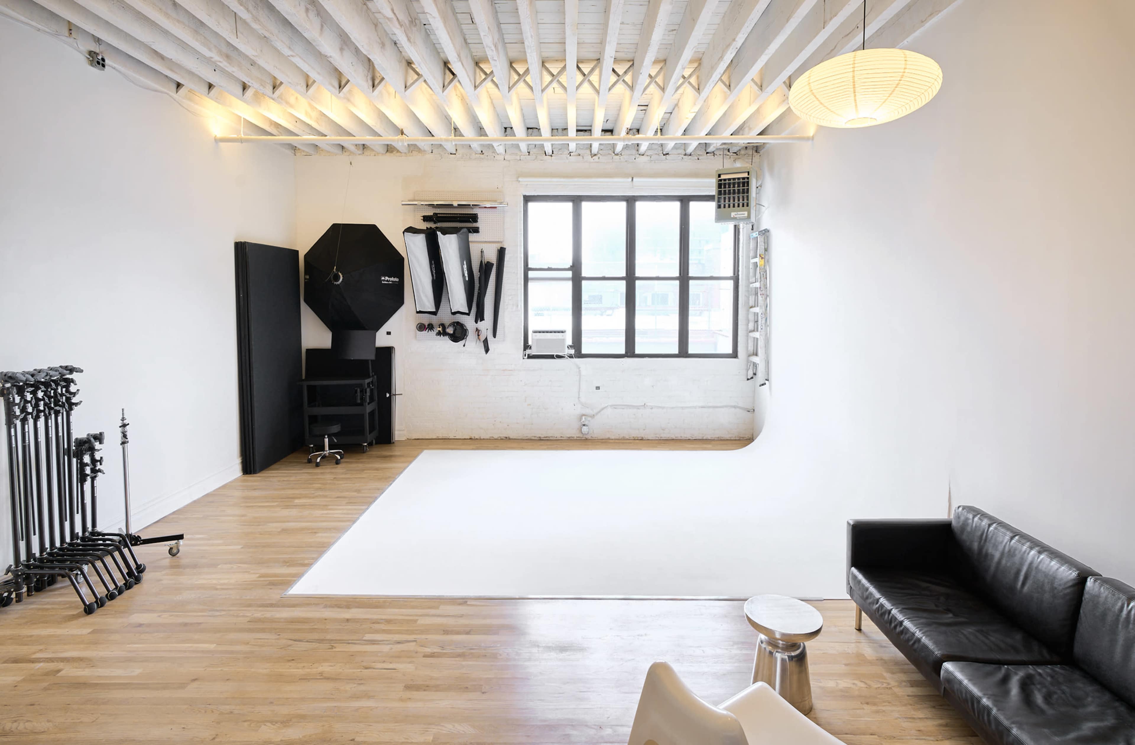 The image shows a spacious, minimalist photography studio with a white backdrop, wooden floors, and a black sofa.