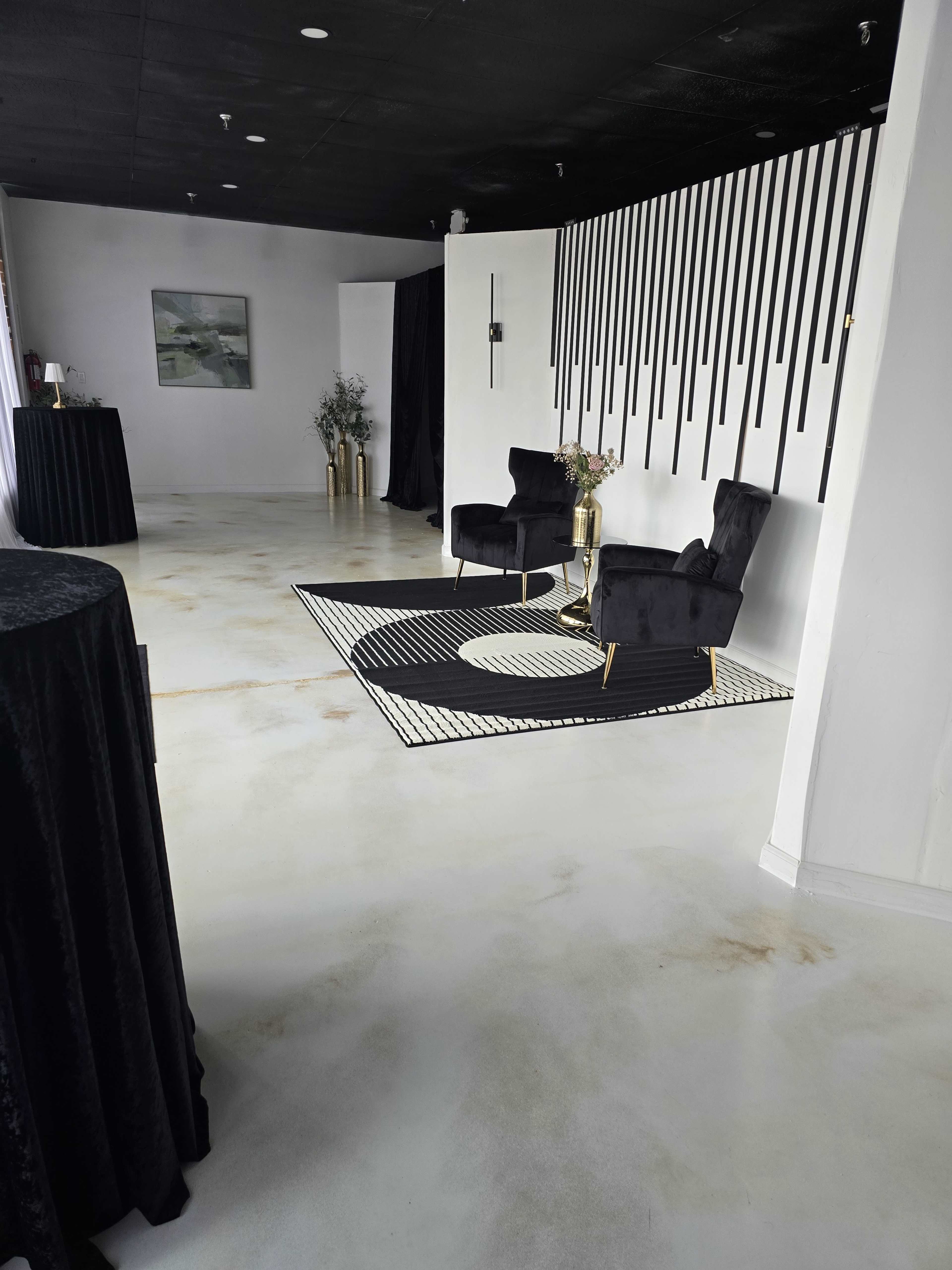 The image shows a modern, minimalist interior featuring two black velvet chairs on a patterned rug, a small gold side table, and decorative elements against a white wall.
