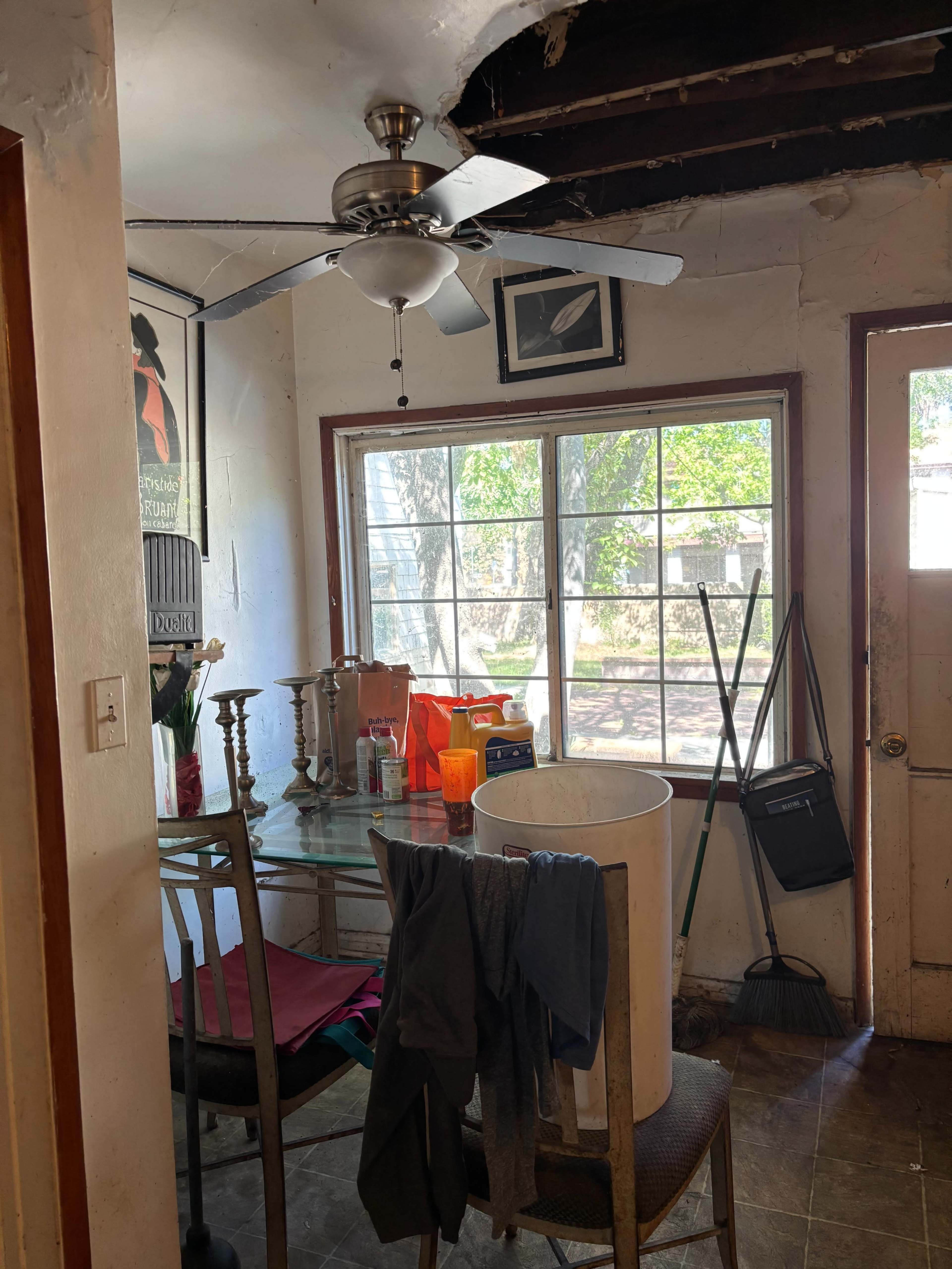 The image shows a small room with a table, chairs, a ceiling fan, and a window overlooking an outdoor area, along with clutter on the table and a door on the right side.