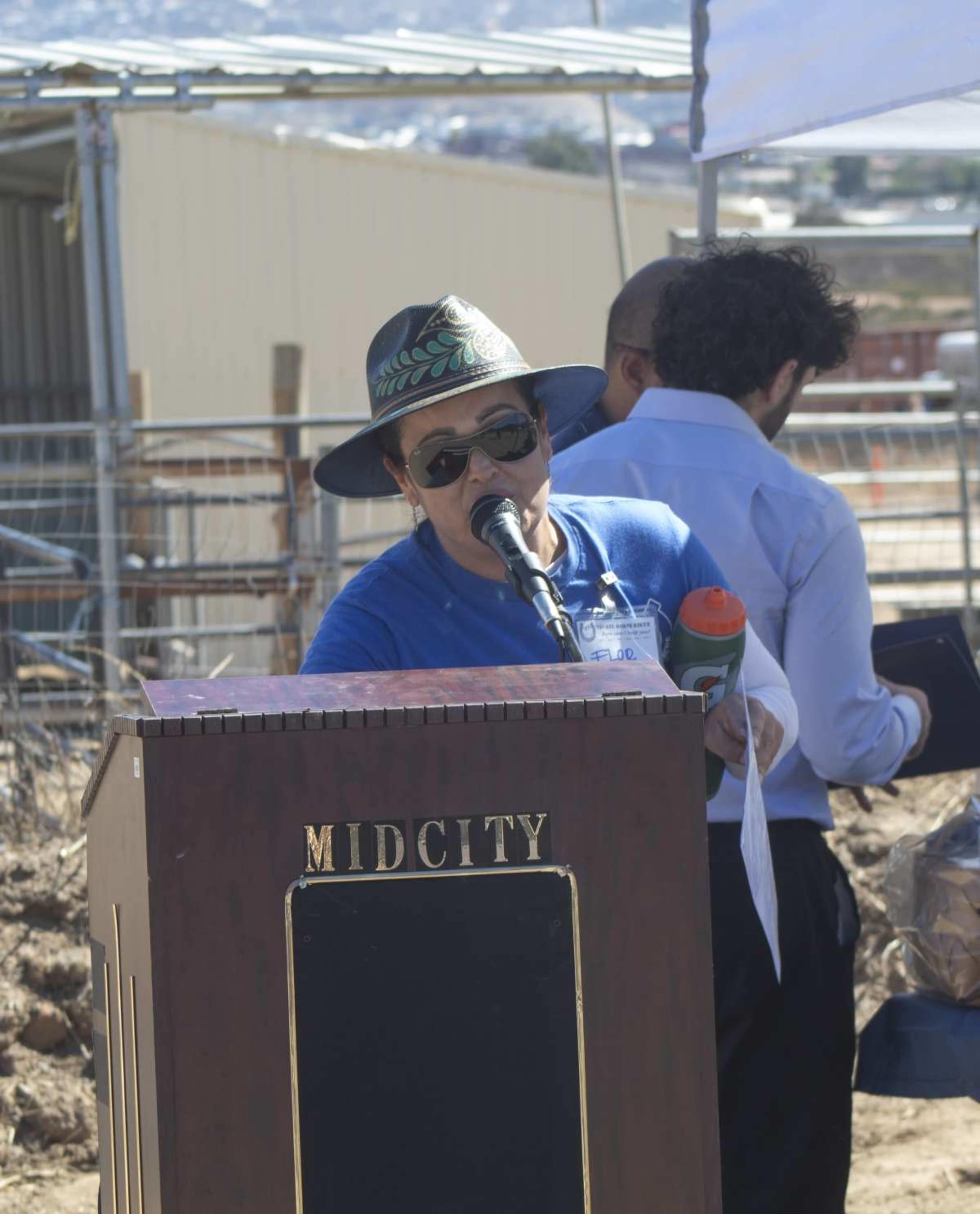 A person in a straw hat speaks at a podium labeled "MIDCITY" during an outdoor event near construction sites.