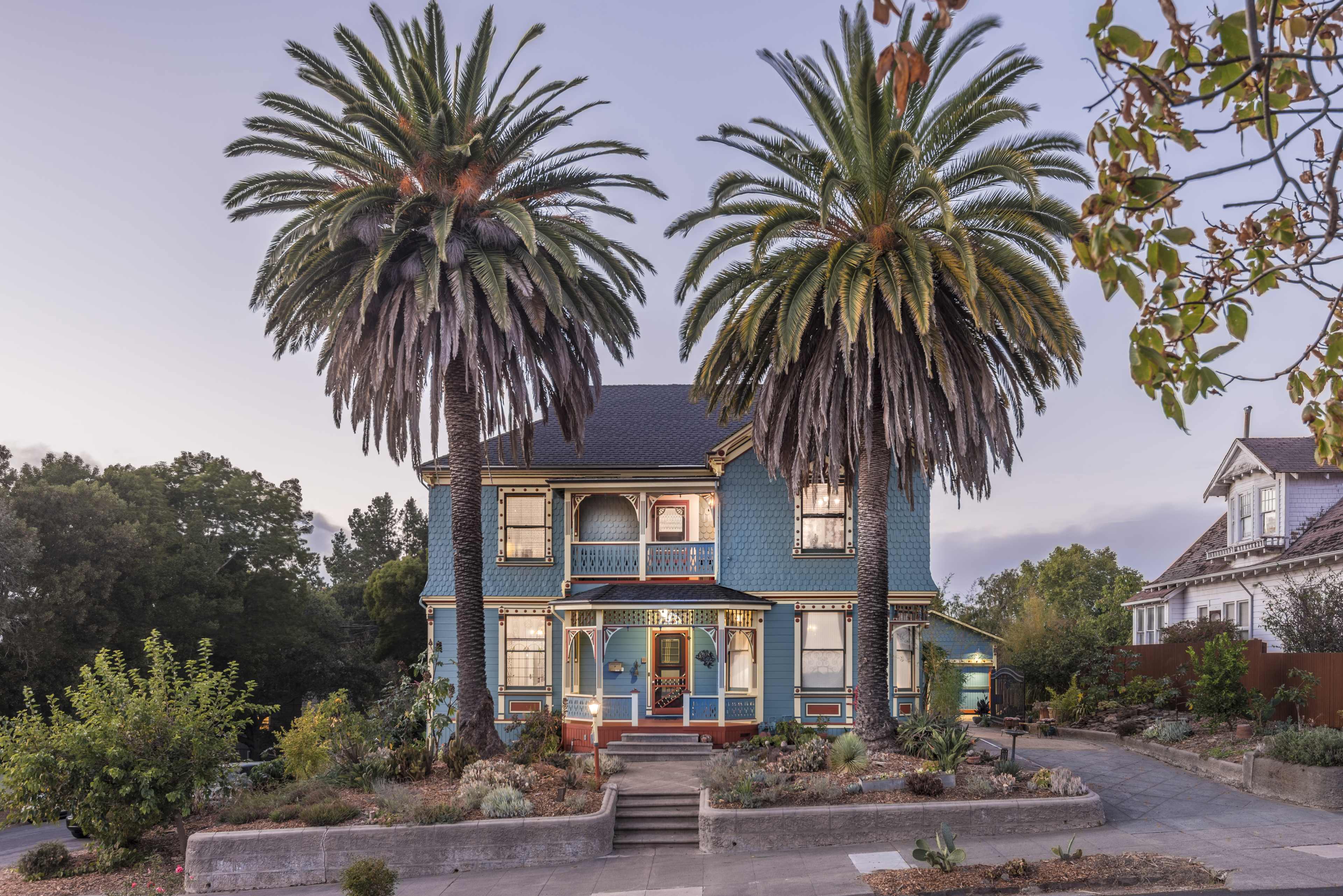 A blue Victorian house with palm trees in front stands in a landscaped yard.
