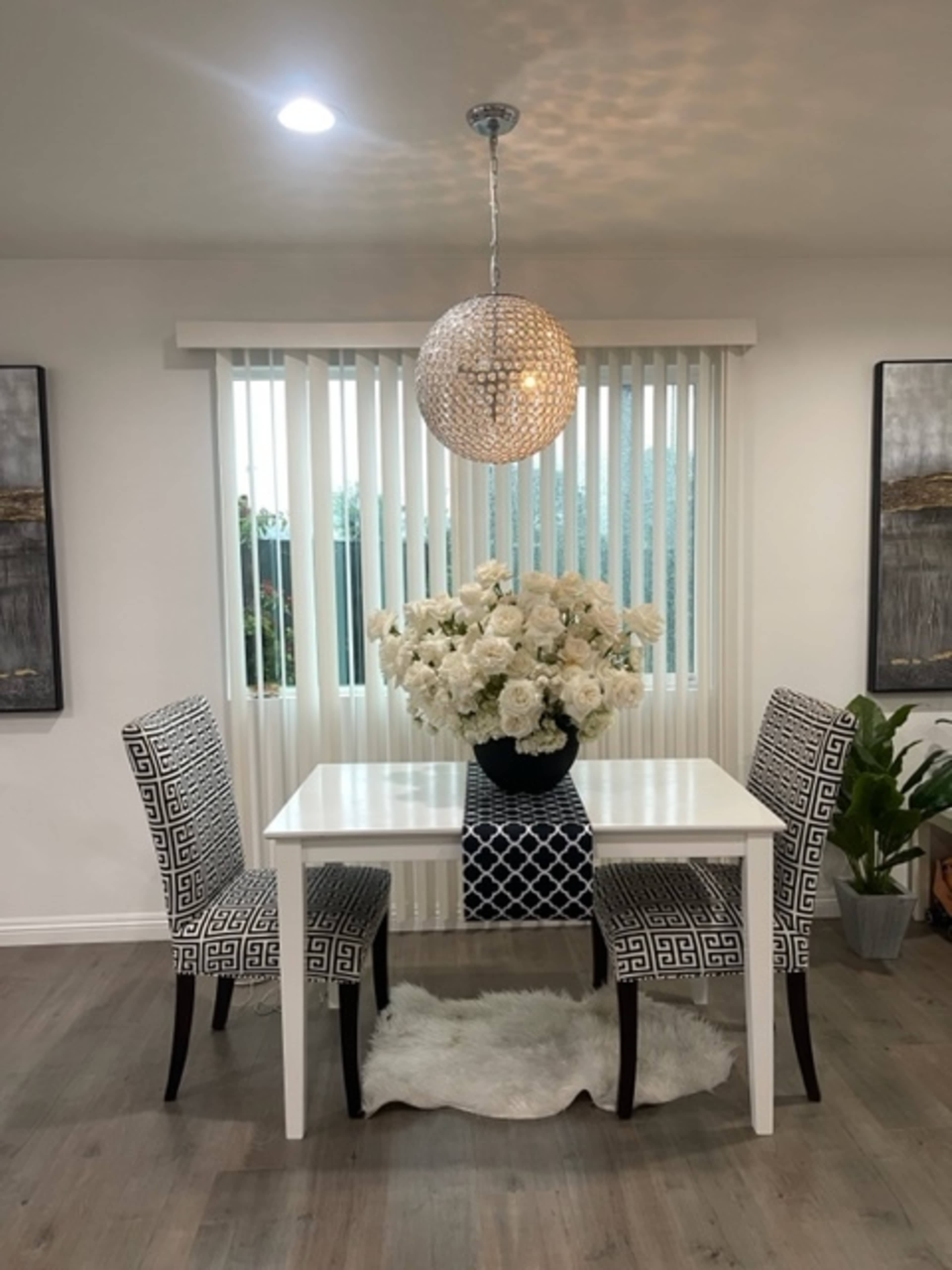 A small dining area features a white table set with two patterned chairs, and a large vase of white flowers under a decorative pendant light.