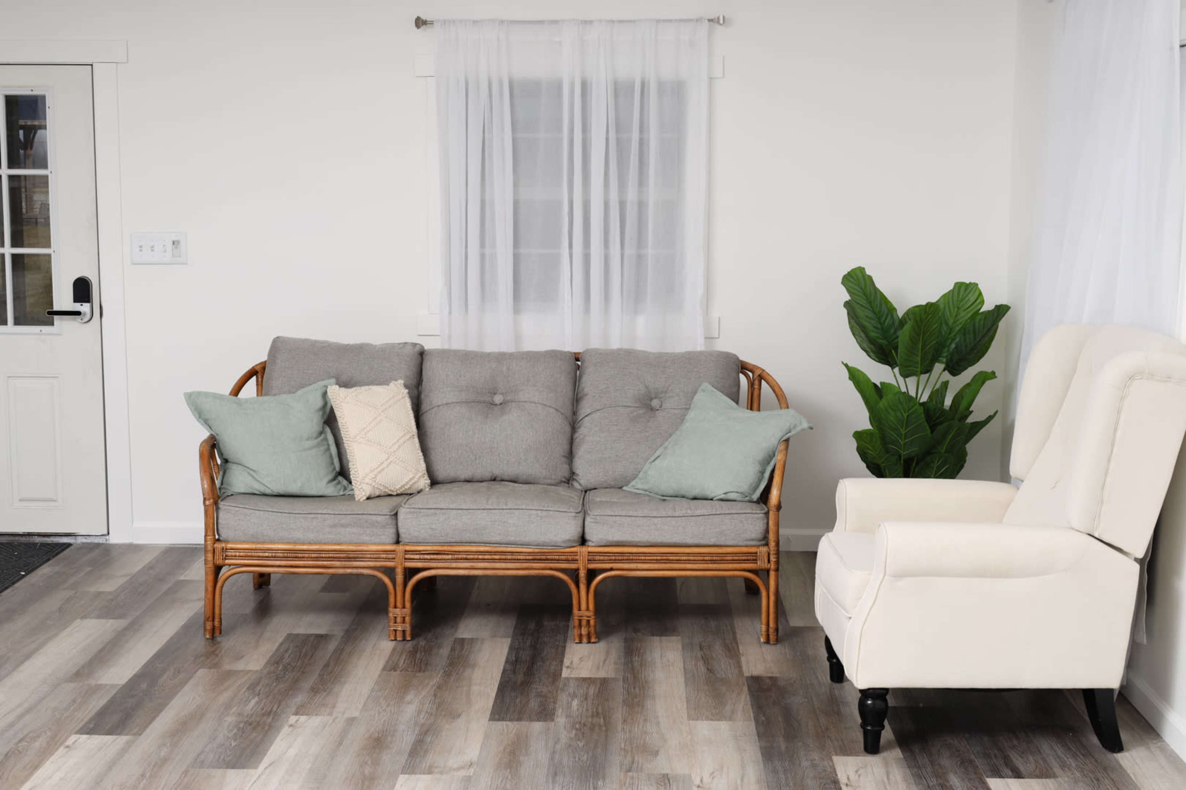 A gray sofa with cushions sits against a white wall, beside a light-colored armchair and a potted plant, with a window and door visible in the background.