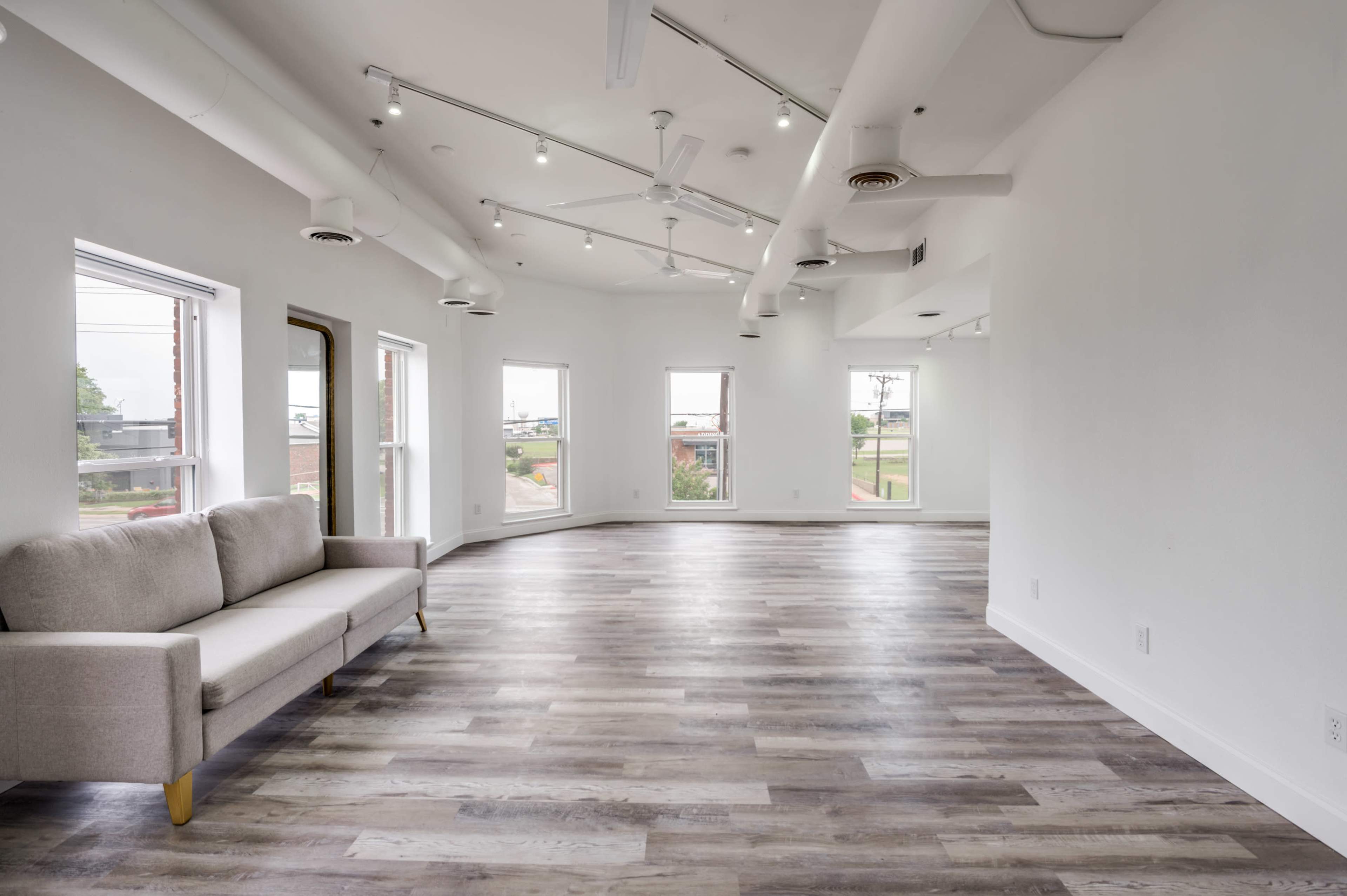 The image shows an empty room with large windows, a light-colored wooden floor, and a couch against one wall.
