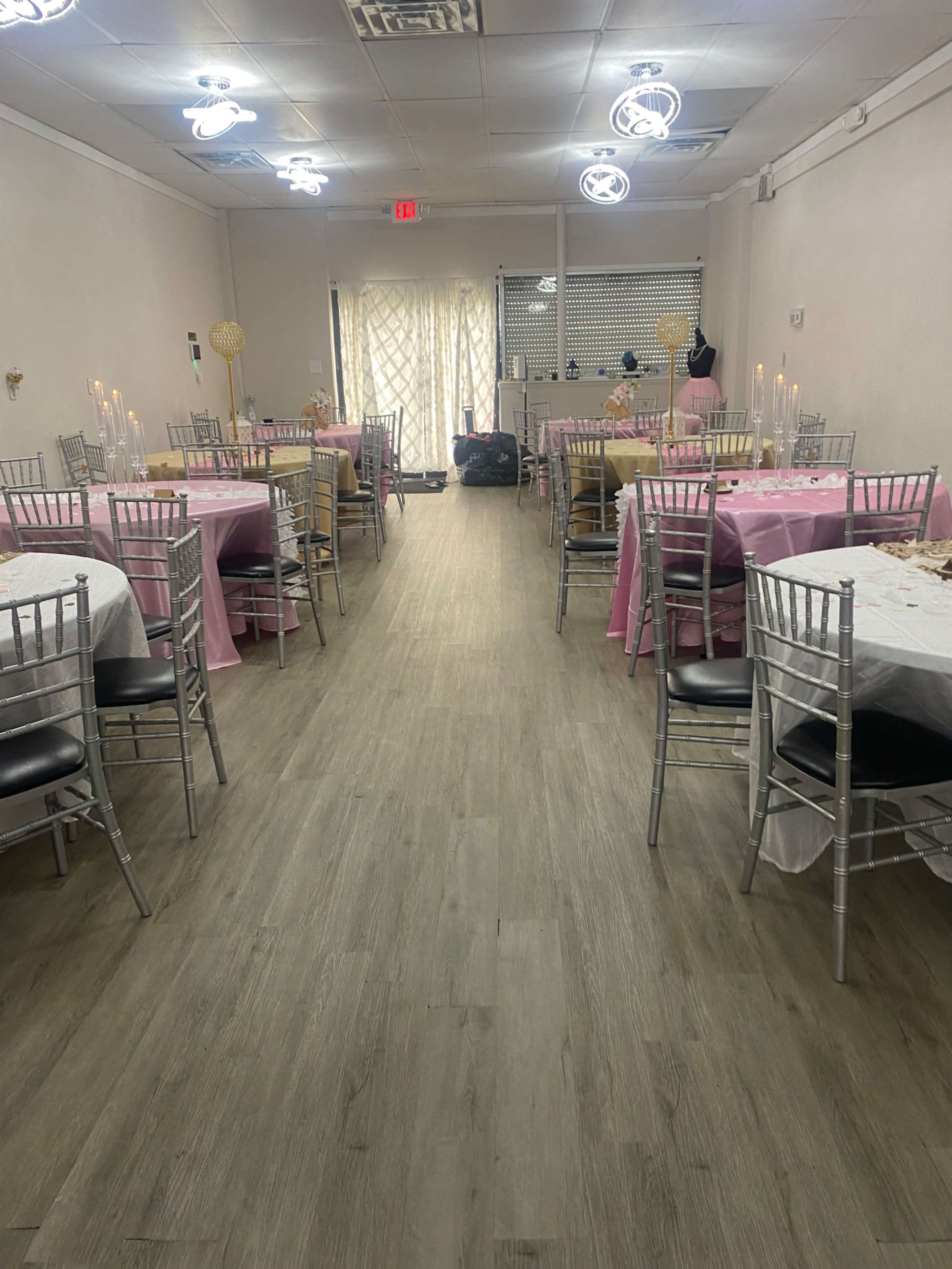 A banquet hall set with tables covered in pink and white tablecloths, lined up along a spacious aisle leading to a doorway at the far end.