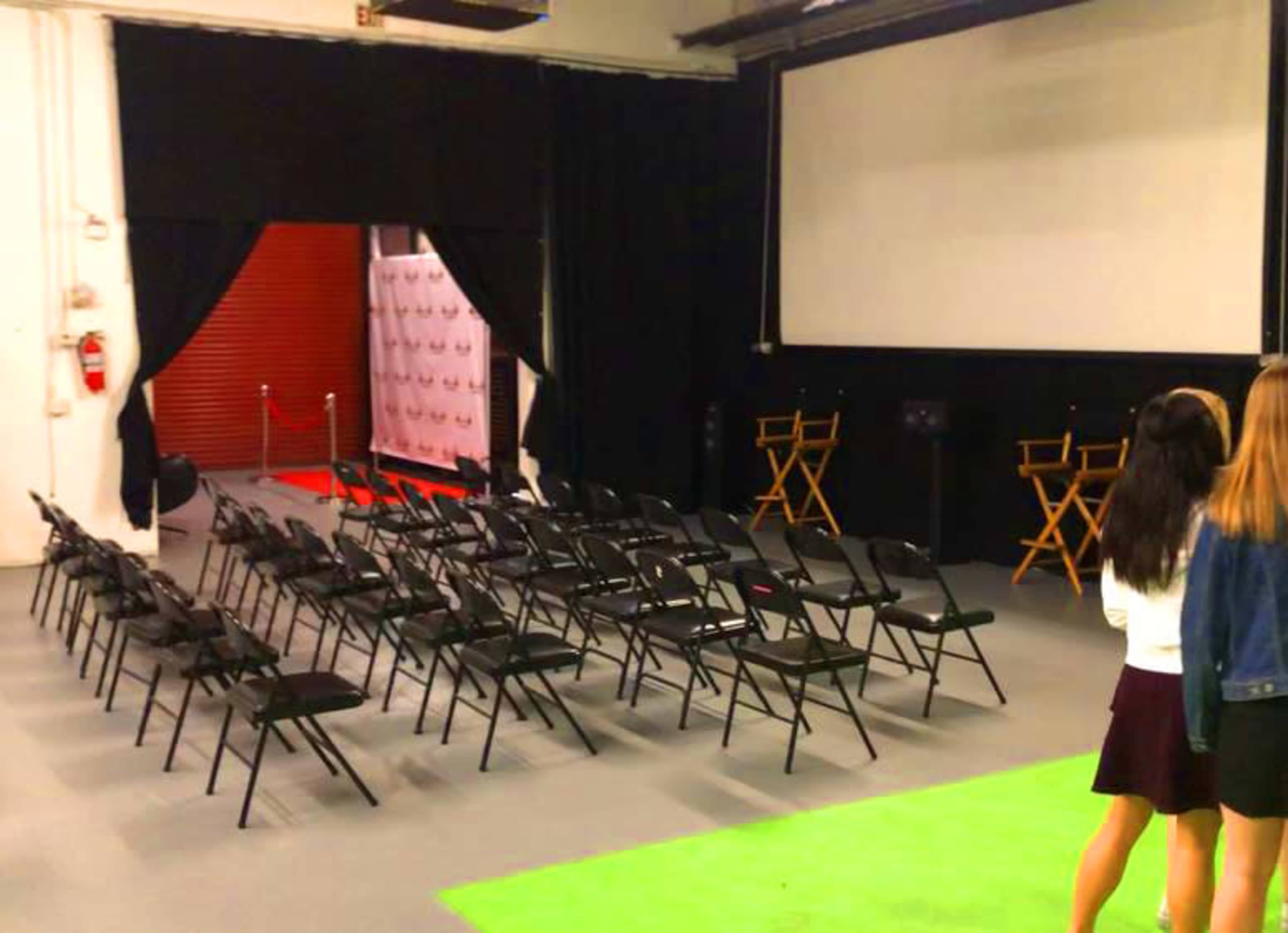 A spacious room features rows of black folding chairs facing a large screen, with a red carpet area and two empty director's chairs in the background.