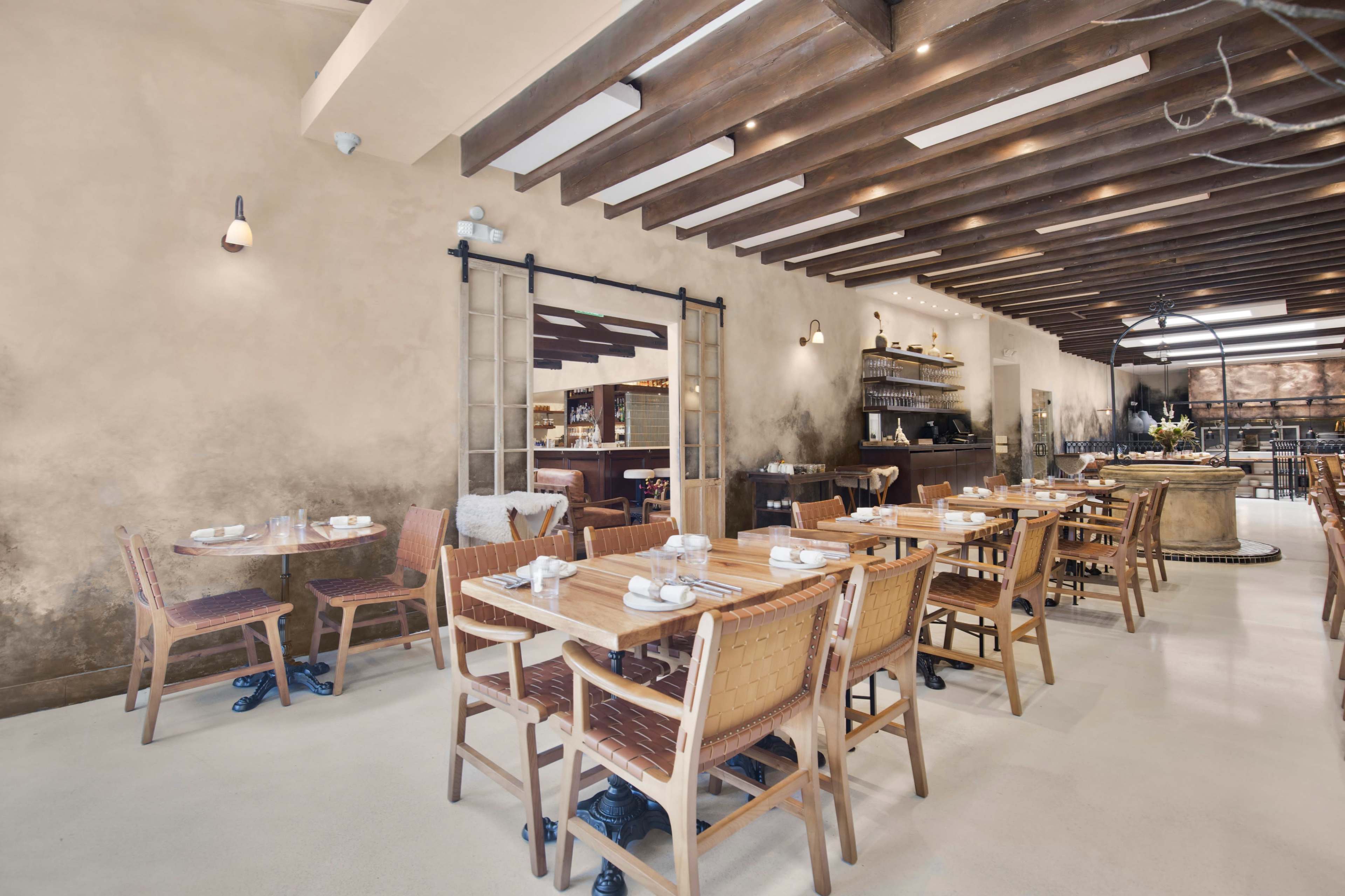 The image shows a spacious restaurant interior with wooden beams, multiple tables set with dinnerware, and an open kitchen area in the background.