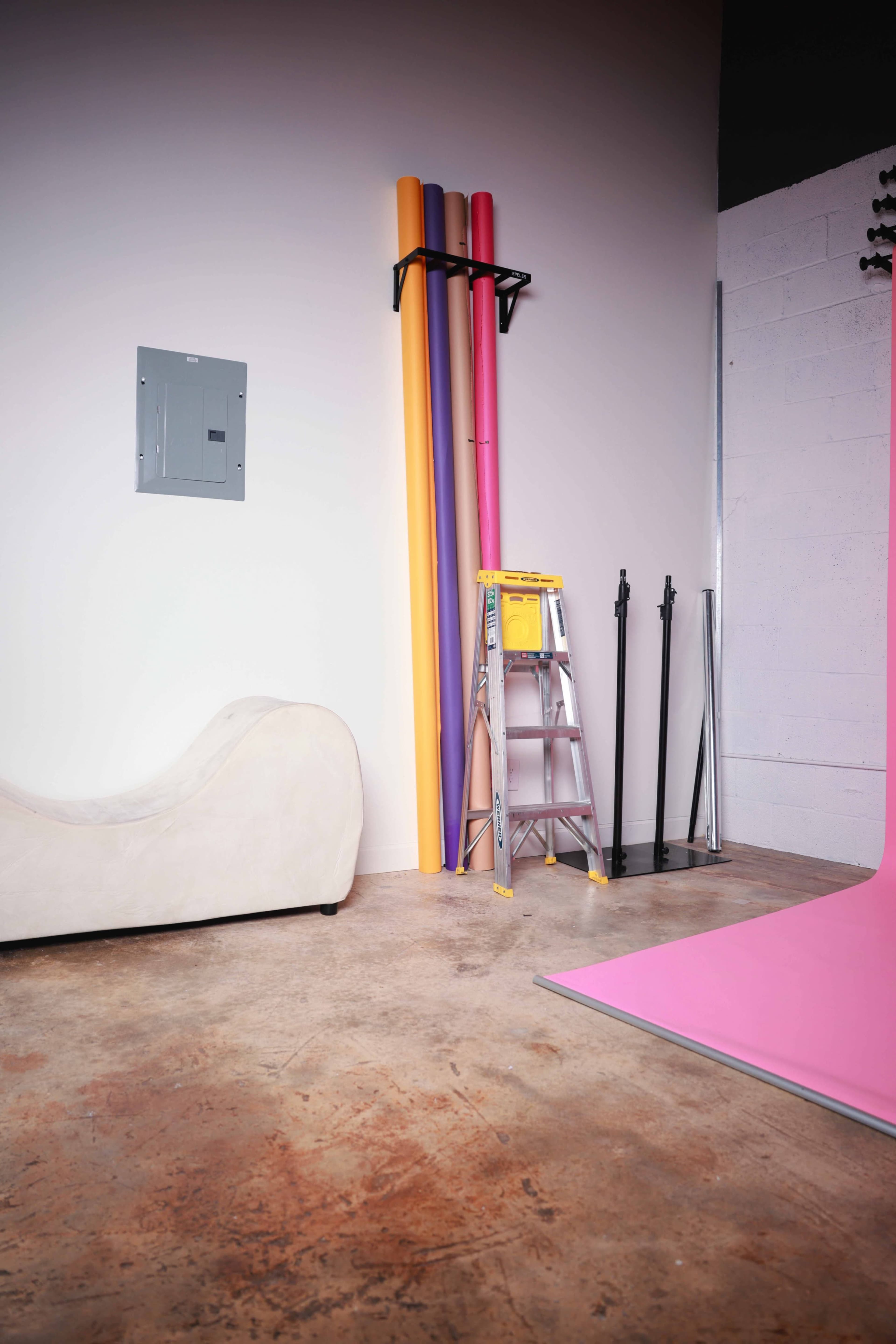 The scene features a beige curved couch, a ladder, and several rolls of colored backdrop paper leaning against a wall in a studio with a concrete floor.